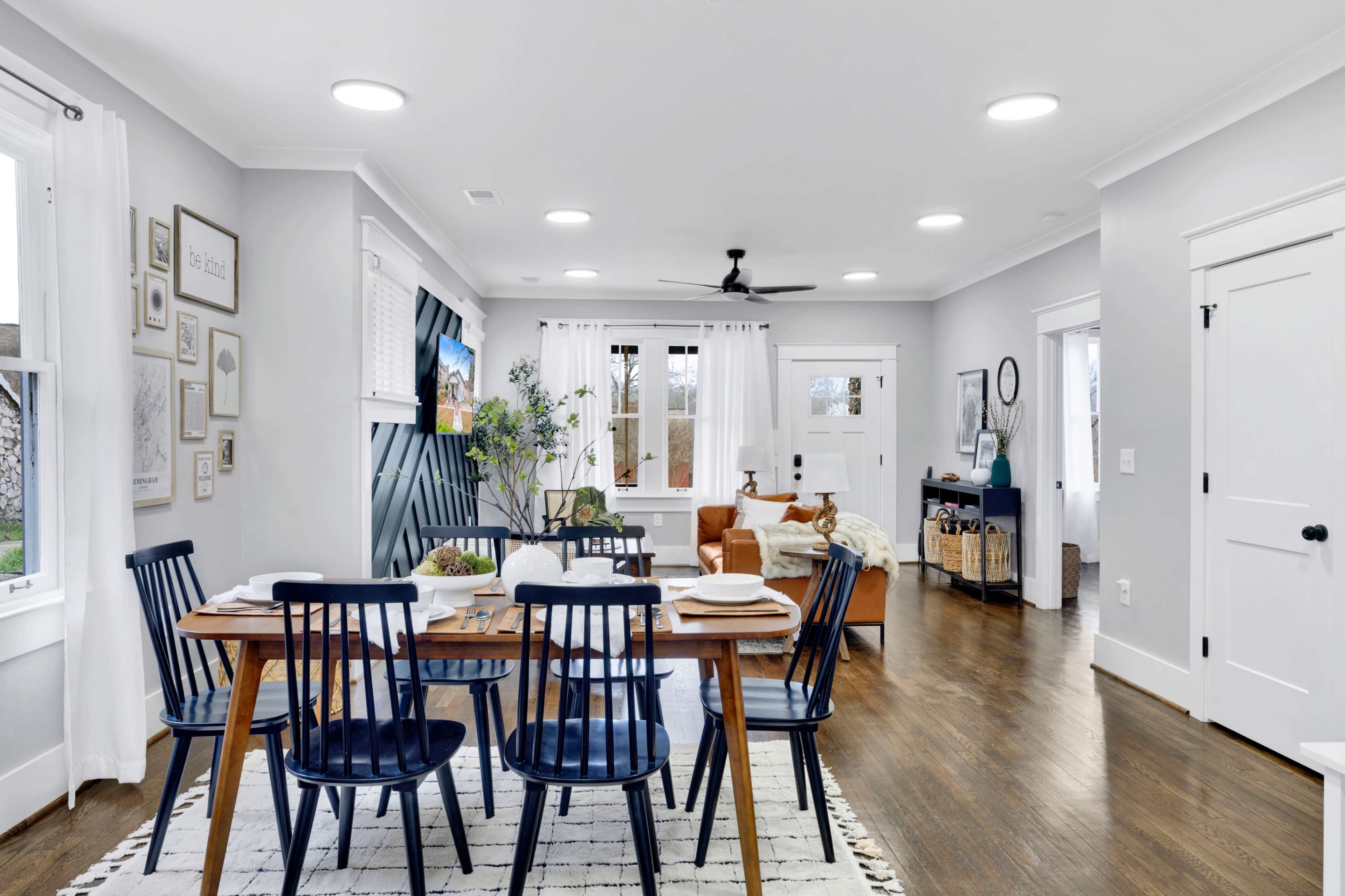 A modern dining area features a wooden table set for meals, surrounded by dark blue chairs, with a living room visible in the background.