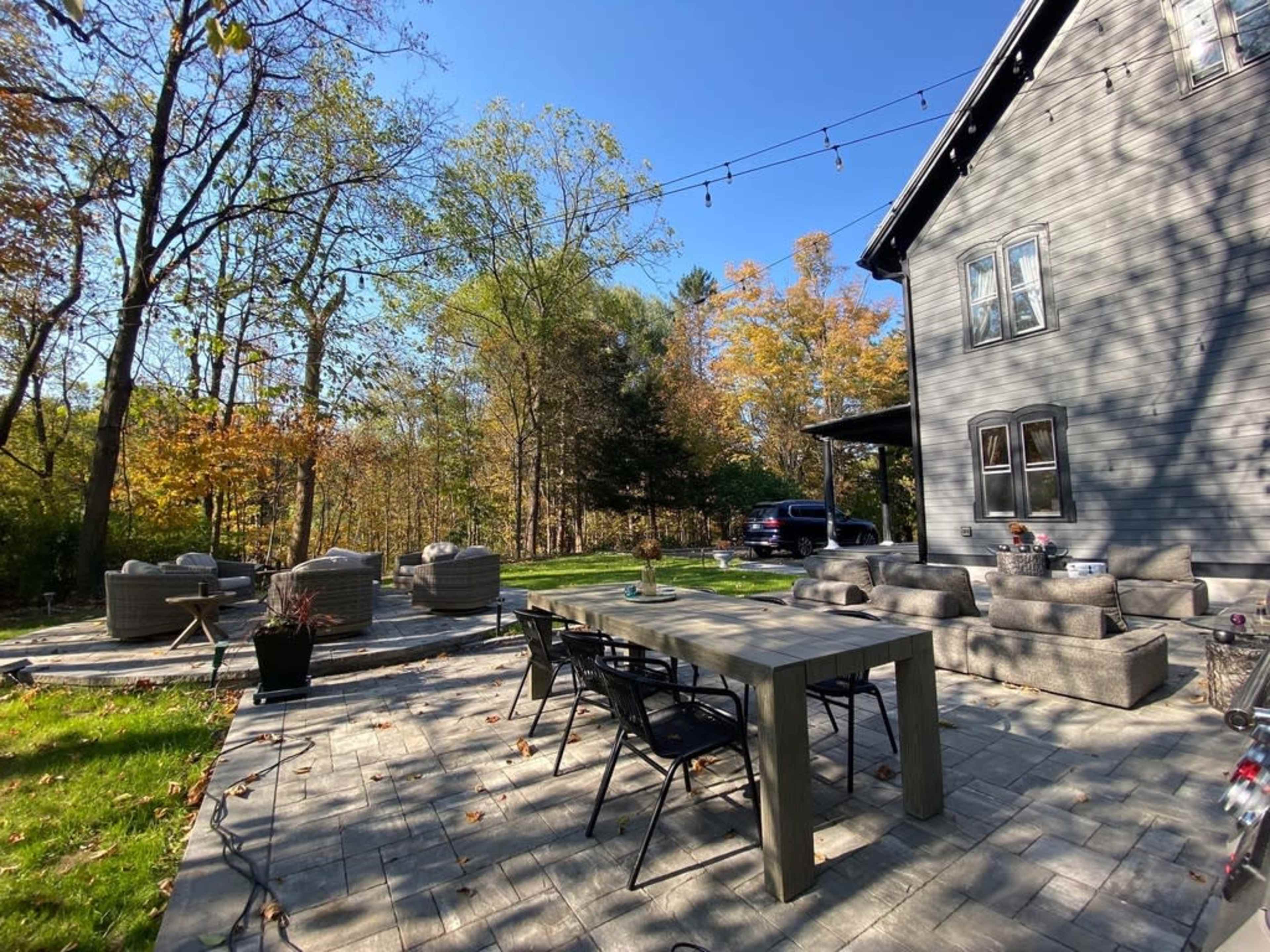 A patio with a rectangular dining table and chairs is set beside a house, surrounded by trees with autumn foliage and arranged seating areas.