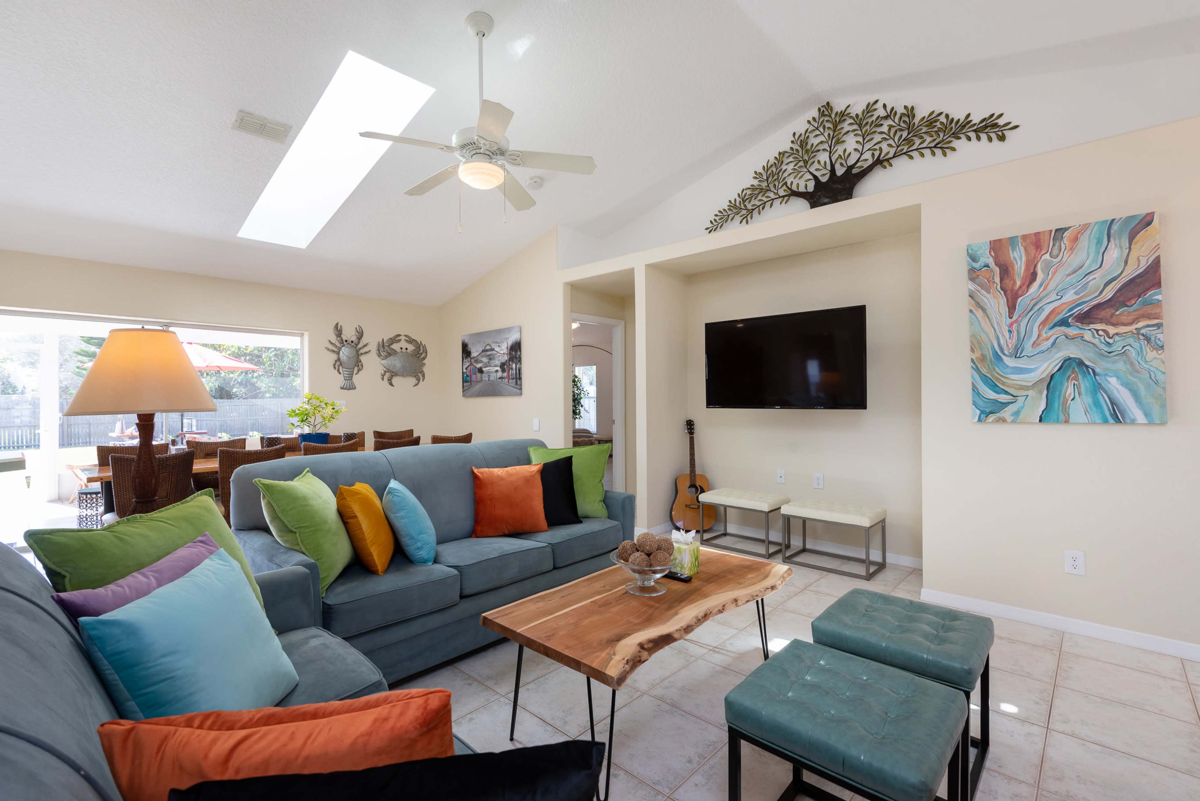 A modern living room featuring a sectional sofa with colorful cushions, a wooden coffee table, and a wall-mounted television, all illuminated by natural light from a skylight.