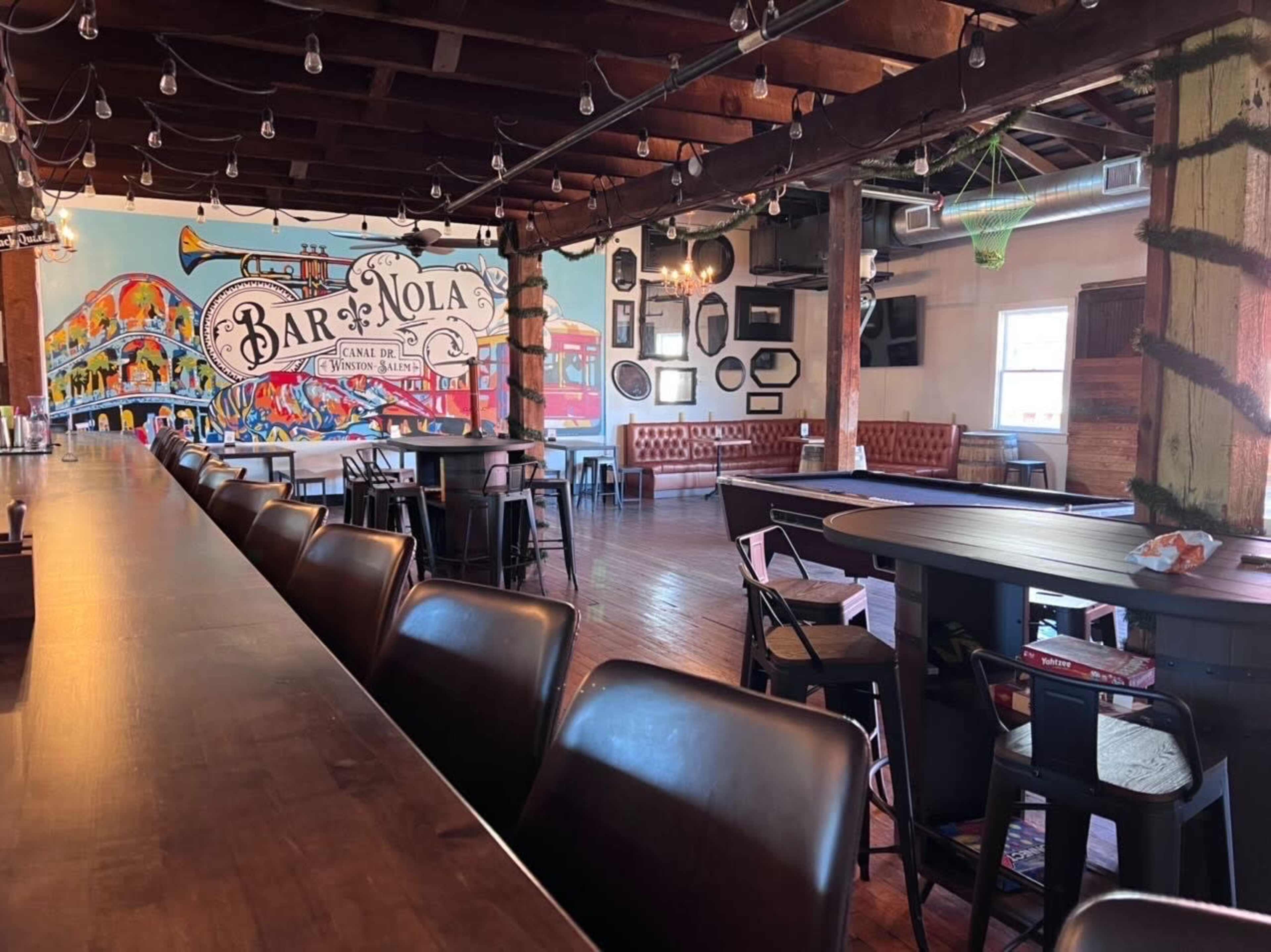 The interior of a bar with a long wooden counter, wooden floors, and colorful murals on the walls.