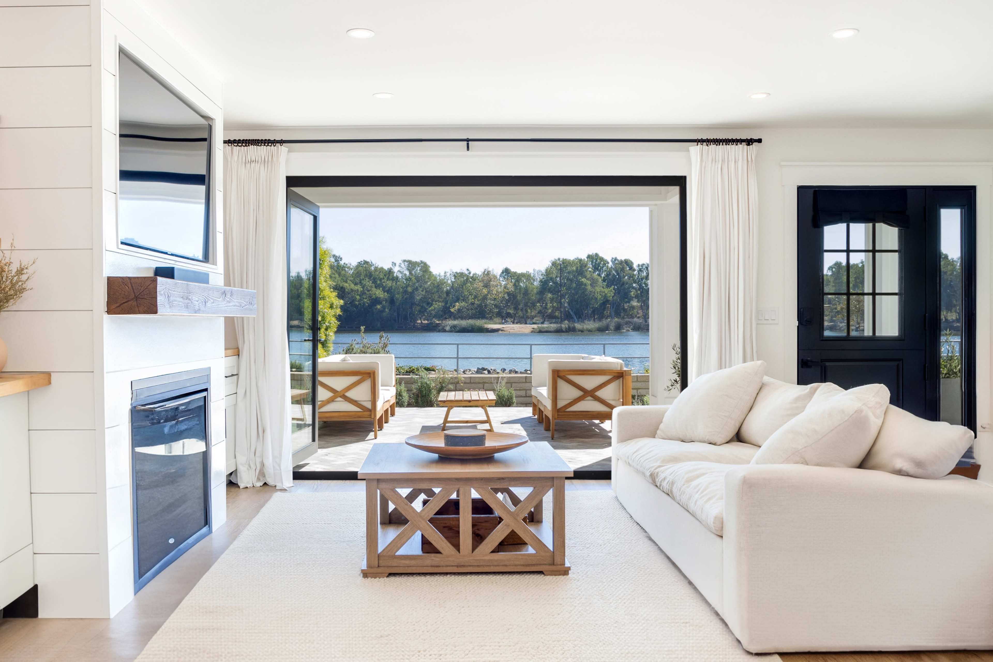 A bright living room with a view of a river, featuring a white sofa, a wooden coffee table, and large glass doors opening to an outdoor seating area.