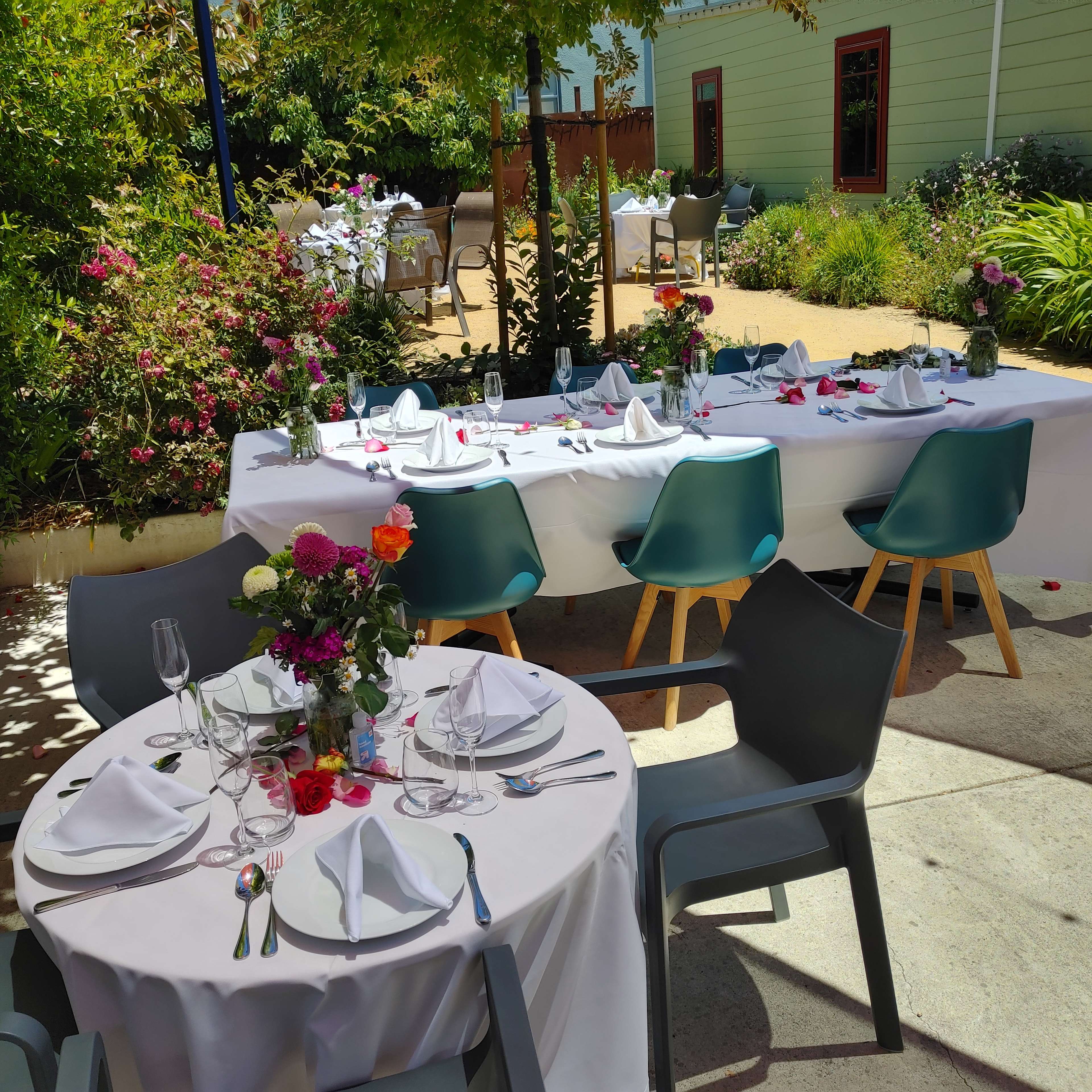 The scene depicts a dining setup in an outdoor area, featuring two tables dressed with white tablecloths and surrounded by green foliage and floral decorations.