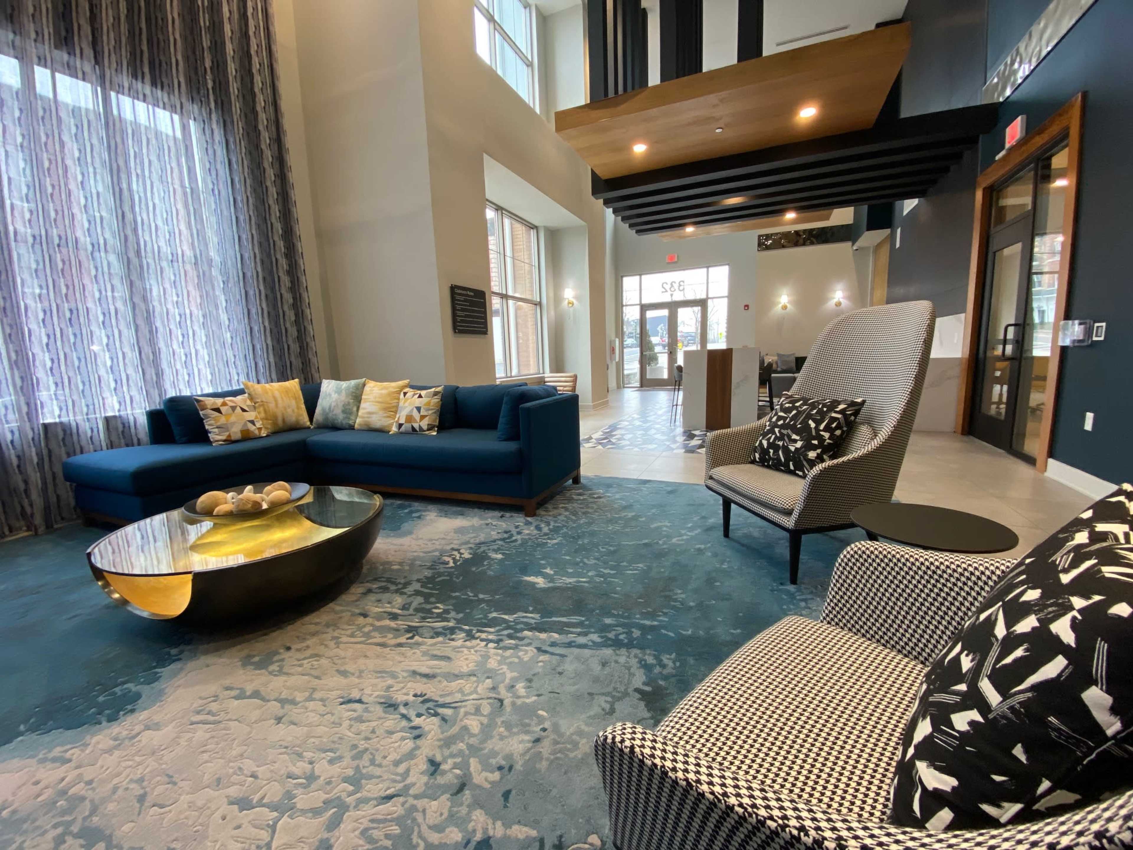The image shows a modern lobby with a large blue sectional sofa, patterned chairs, a round coffee table, and tall windows allowing natural light to enter.