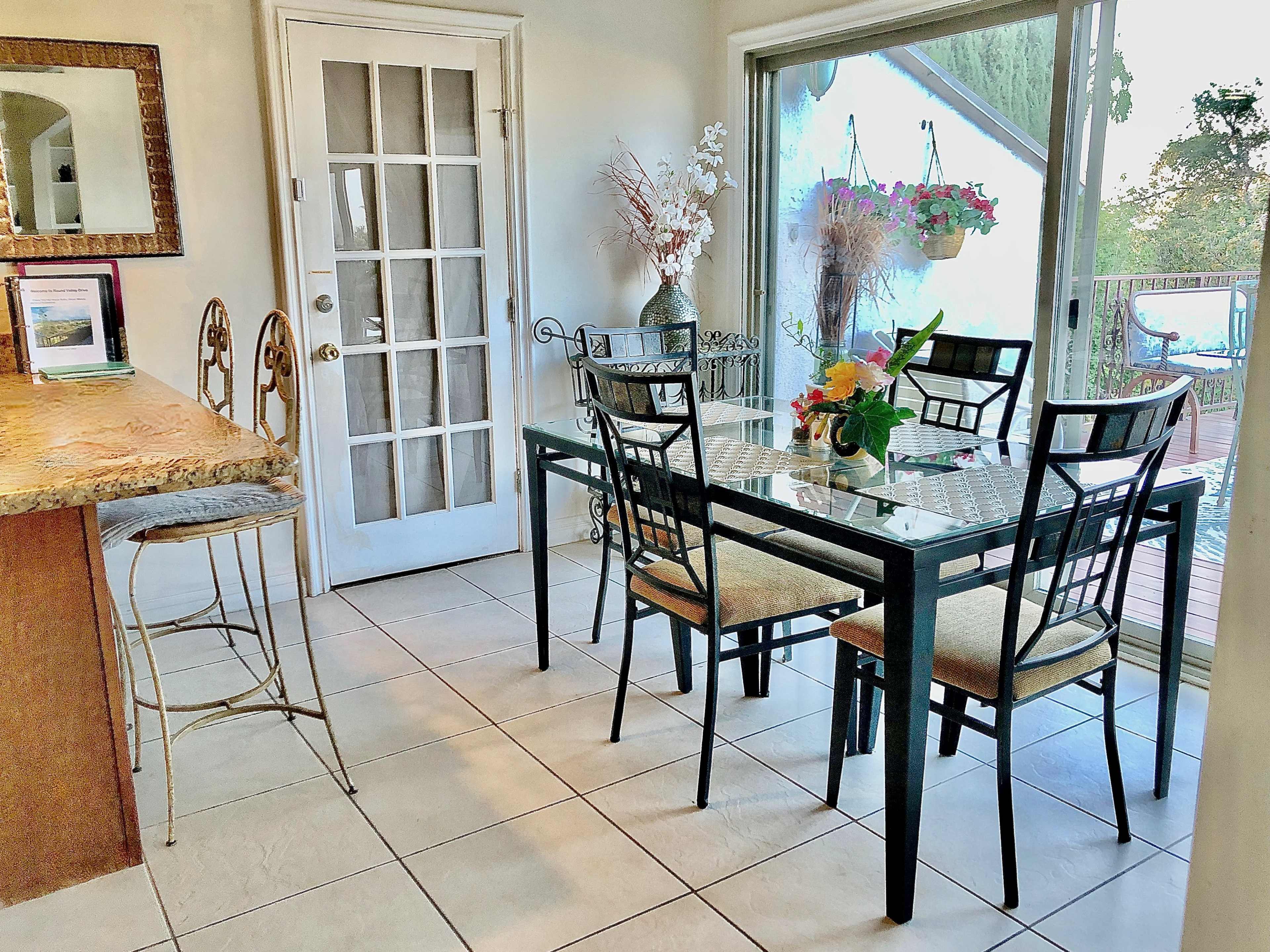 A glass-top dining table with four chairs is positioned near a sliding glass door that leads to an outdoor deck, with a small vase of flowers on the table.