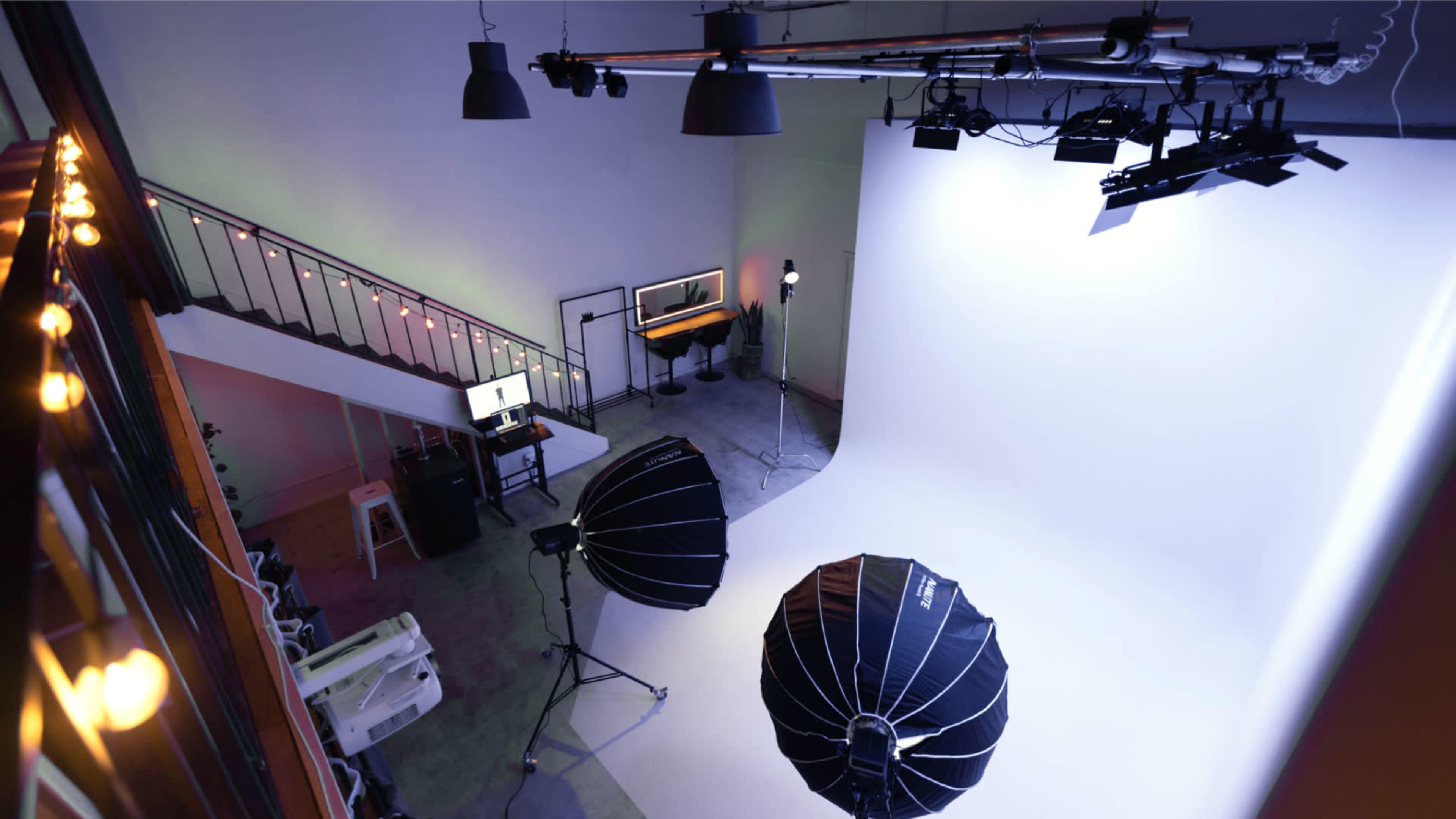 A photography studio features a large white backdrop, lighting equipment, and a workspace with a computer and seating on an upper level.