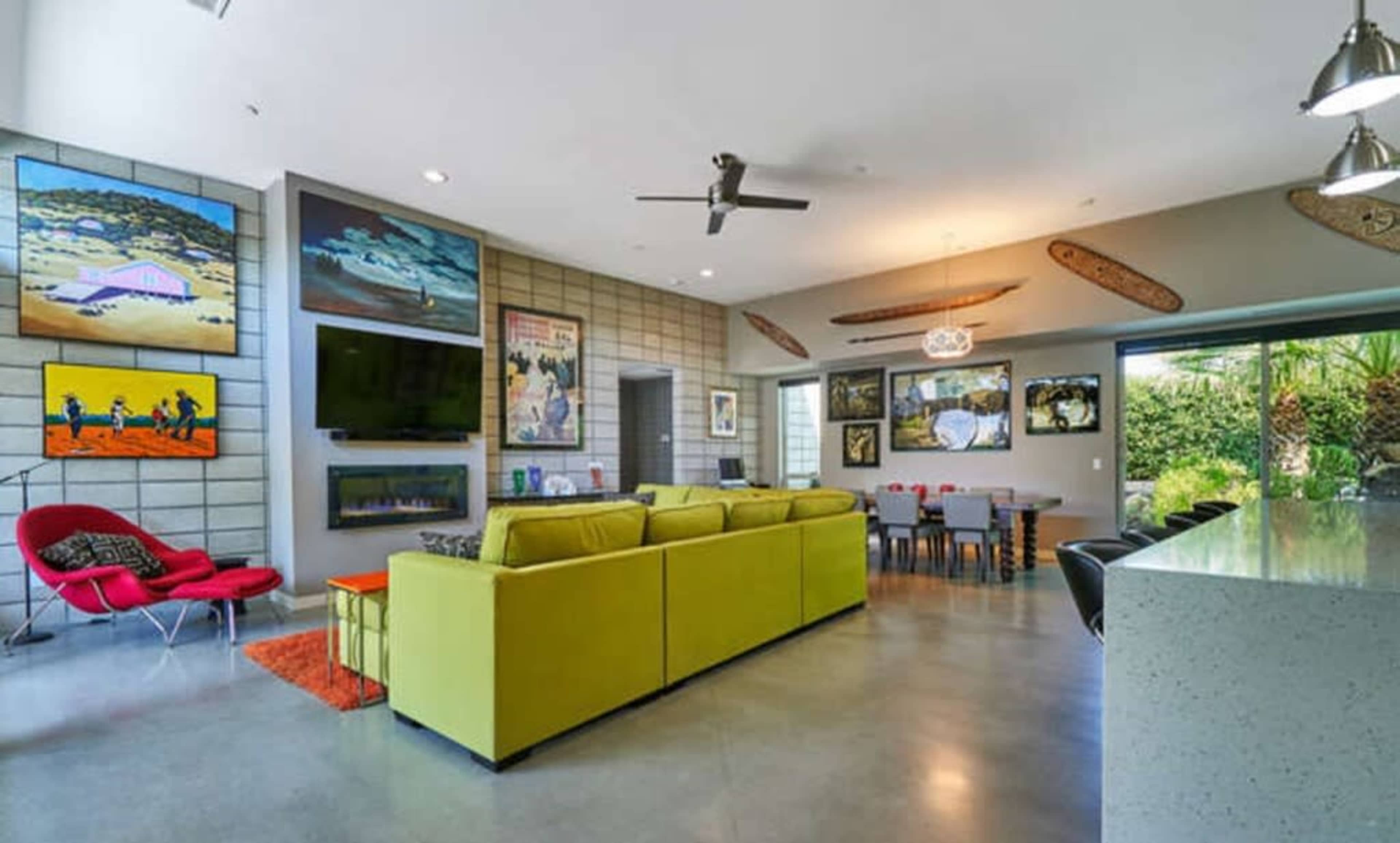 The room features a modern living space with a green sectional sofa, an entertainment area with flat-screen TVs, and colorful artwork on the walls, leading to an outdoor view through large windows.