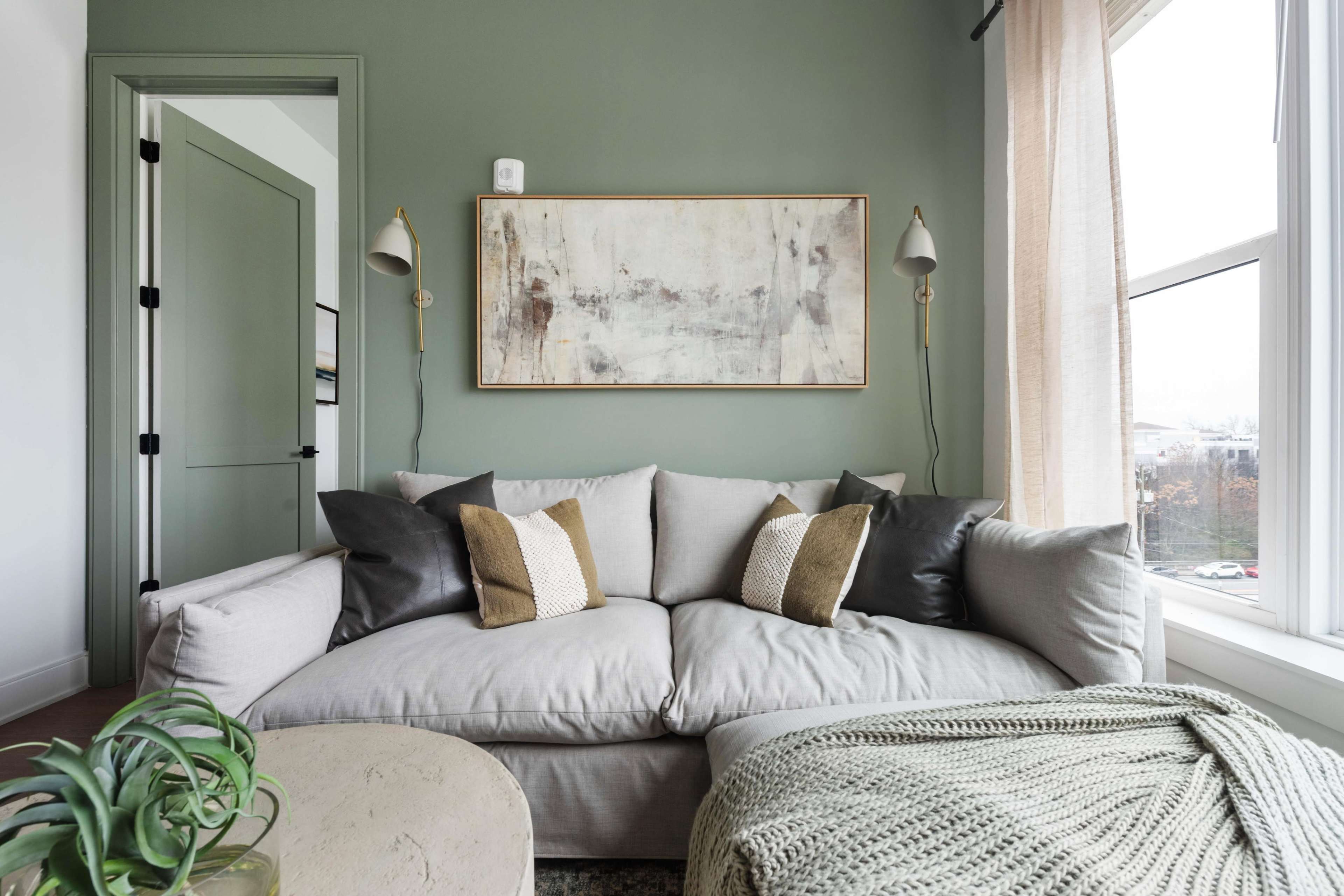 A comfortable light gray sofa with decorative pillows is positioned in a living room featuring pale green walls and large windows.
