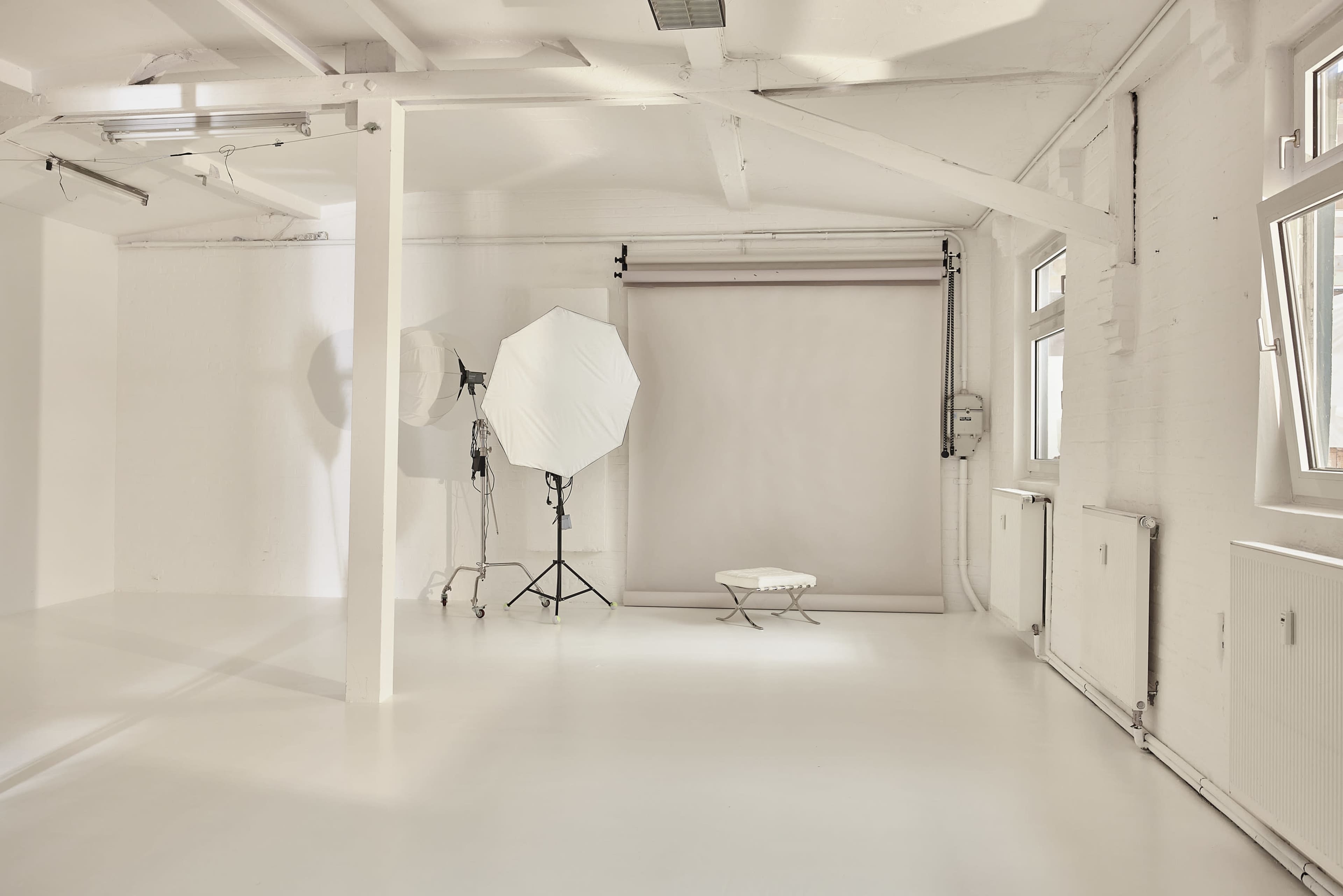 A minimalistic studio space features a white backdrop, a light stand with a large umbrella diffuser, and a small white furniture piece.