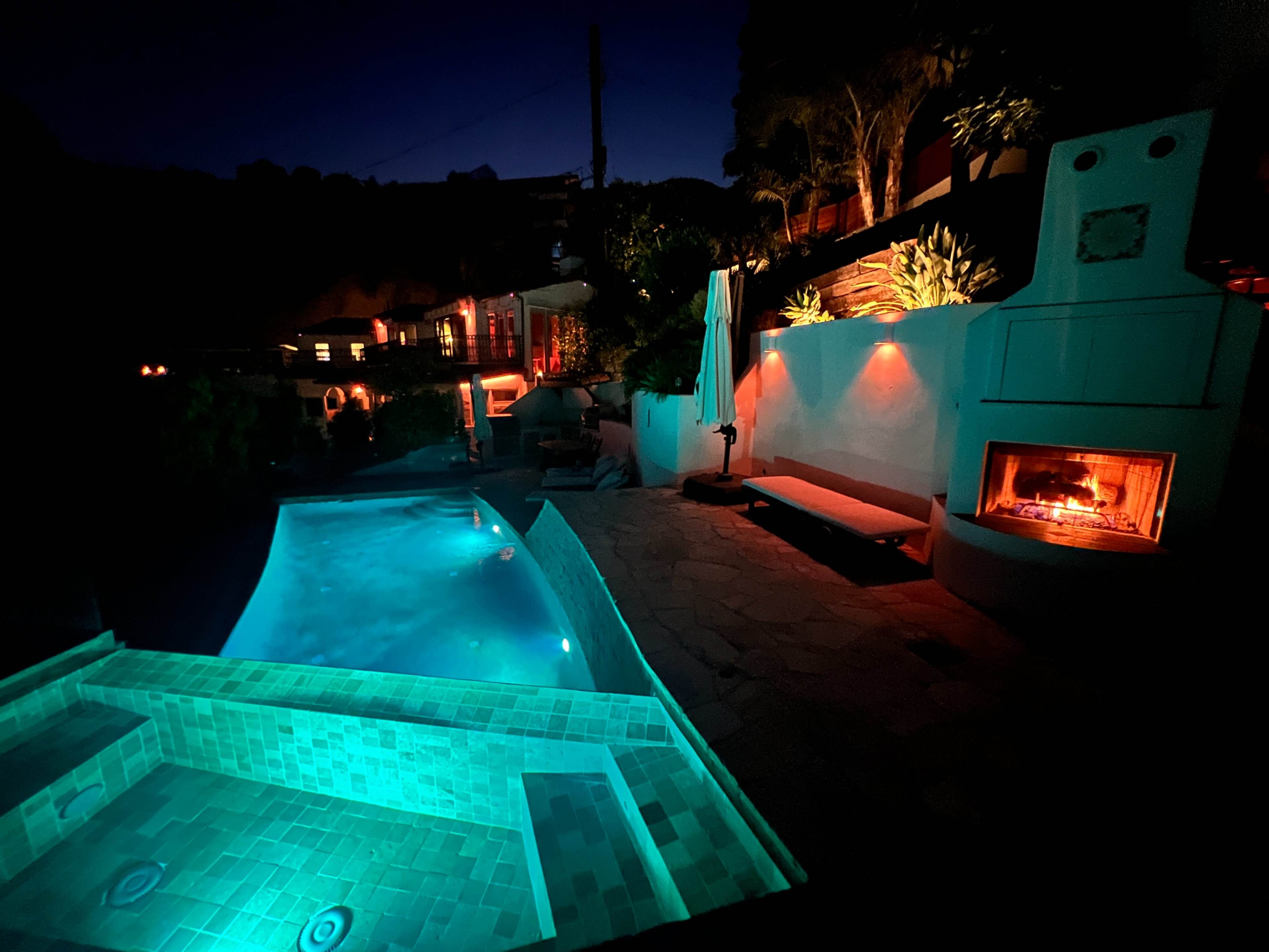 The image shows a backyard pool area illuminated at night, featuring a spa, a swimming pool, a fireplace, and landscaped walls.