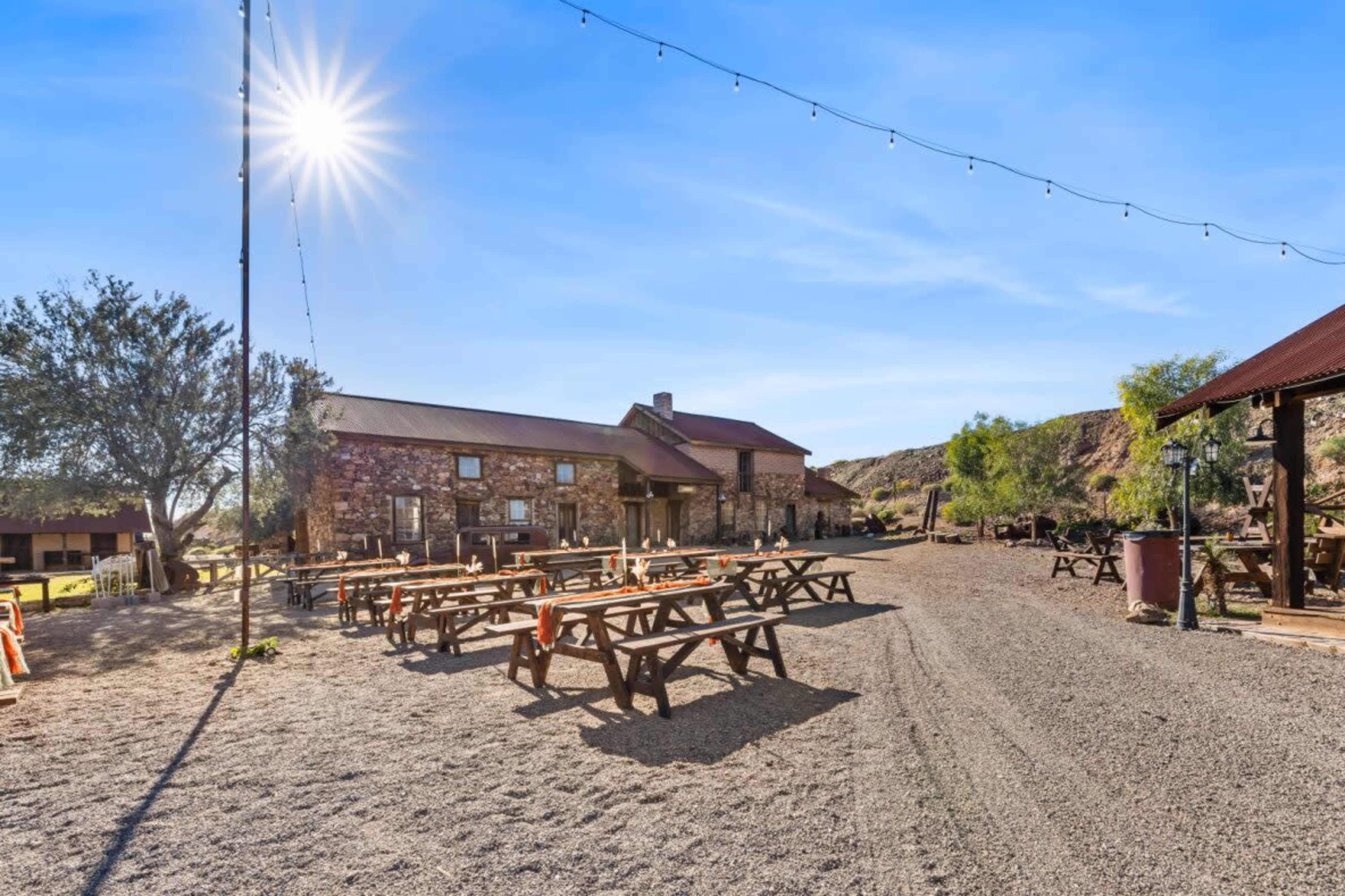 The scene features a gravel pathway lined with wooden picnic tables under a clear sky, with a building made of stone in the background and string lights overhead.