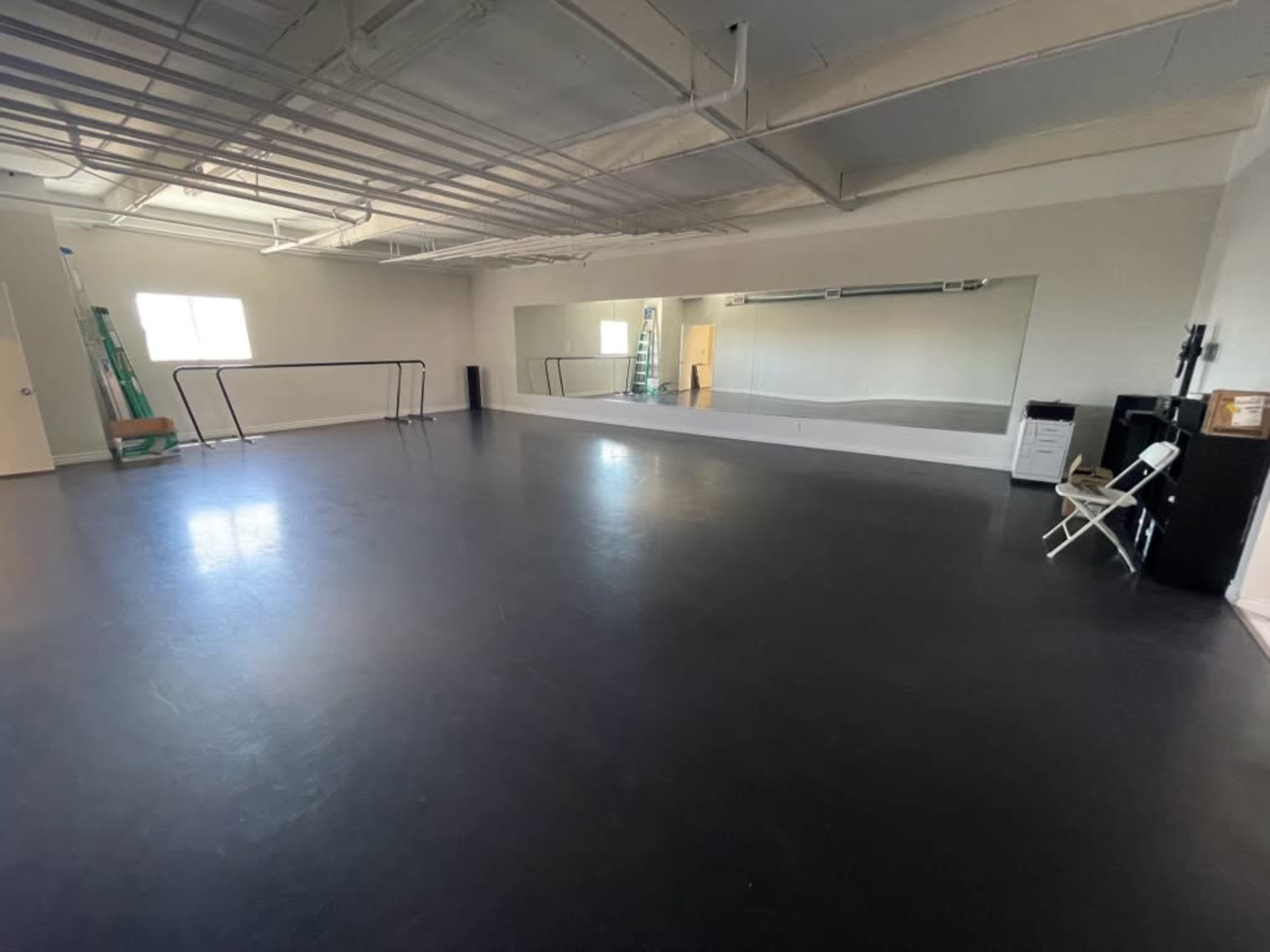 An empty dance studio features a mirrored wall, a black floor, and a folding chair along with a ladder and black equipment in the corner.