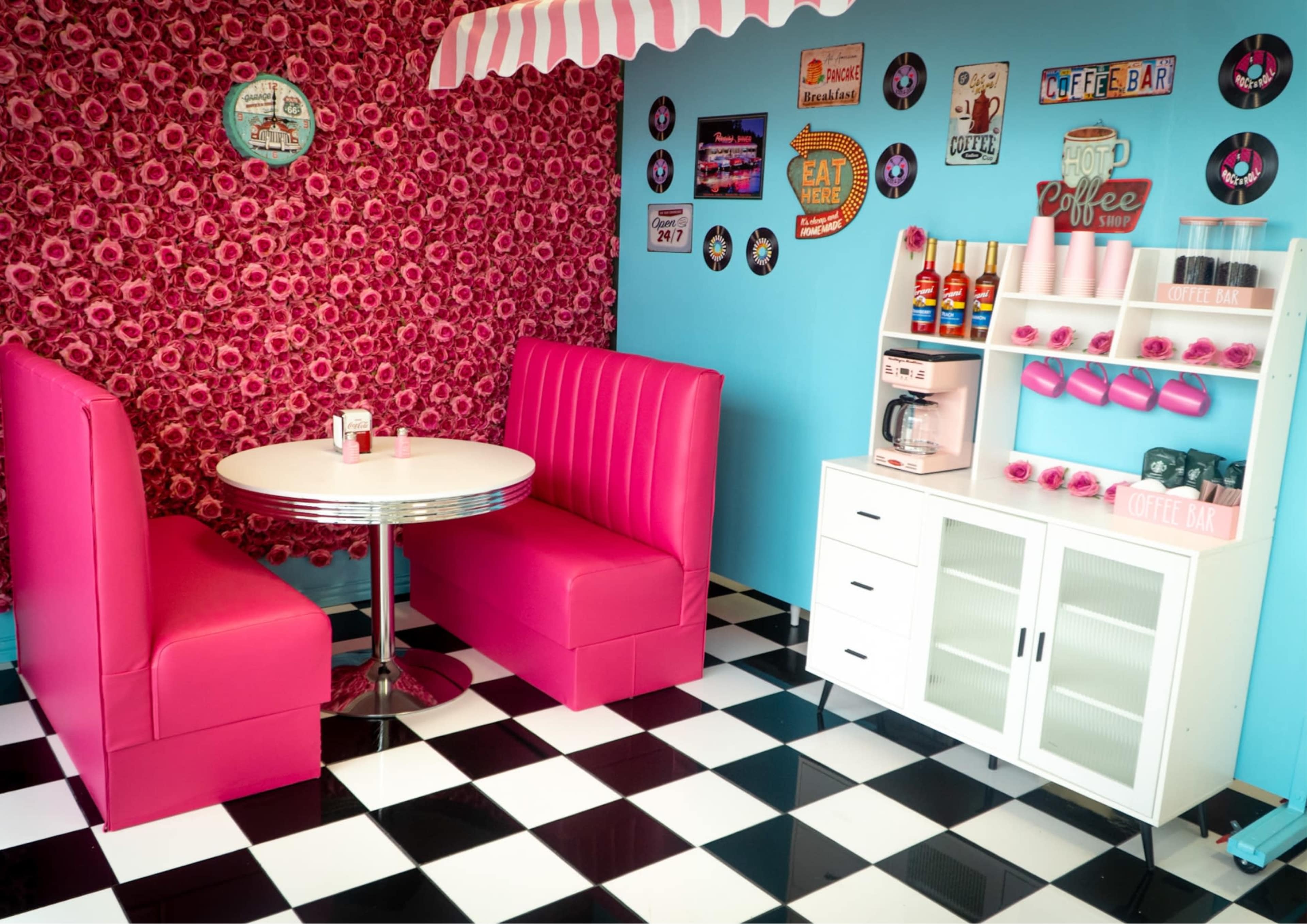 The image depicts a retro-style diner interior with pink booths, a circular white table, and a decorative wall featuring a floral pattern and various retro signs.
