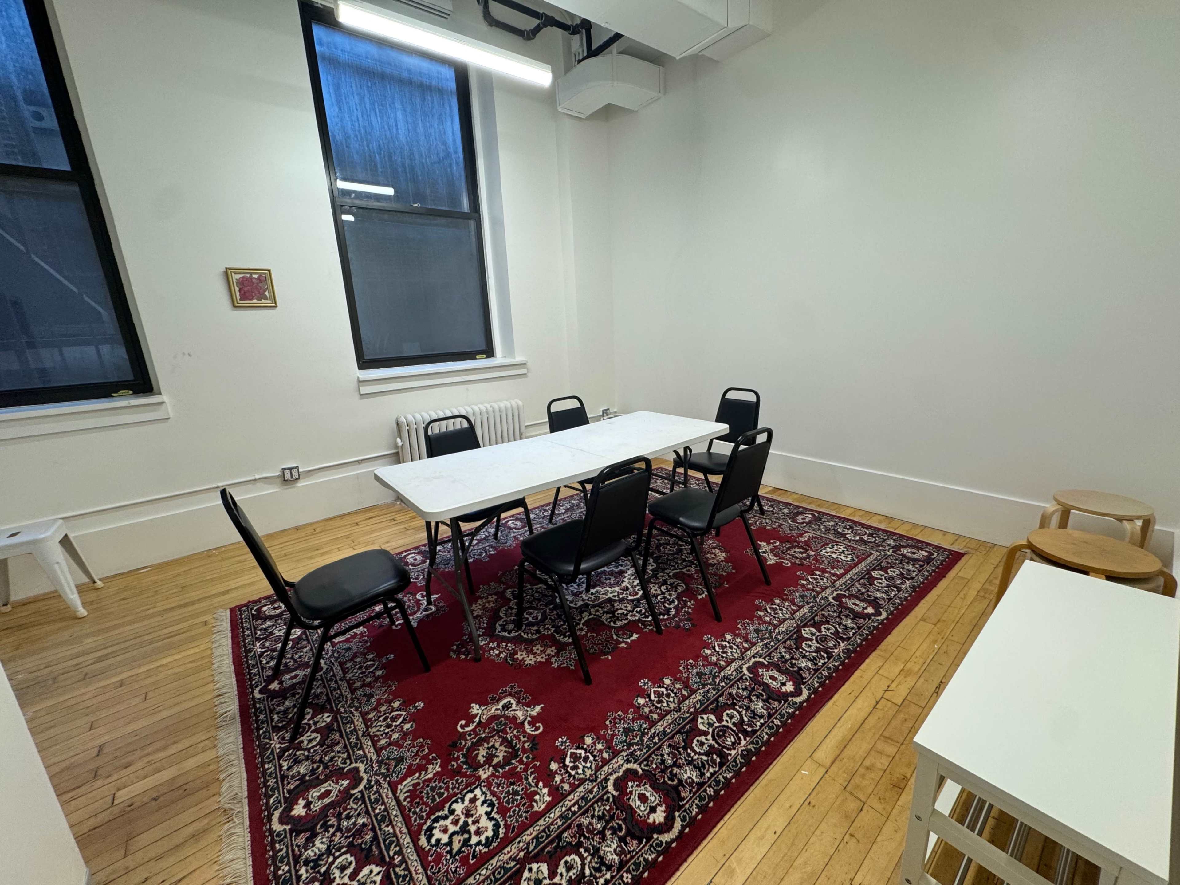 A sparsely furnished meeting room features a rectangular table surrounded by six black chairs, with a red area rug on the wooden floor and a few other small furniture pieces against the walls.