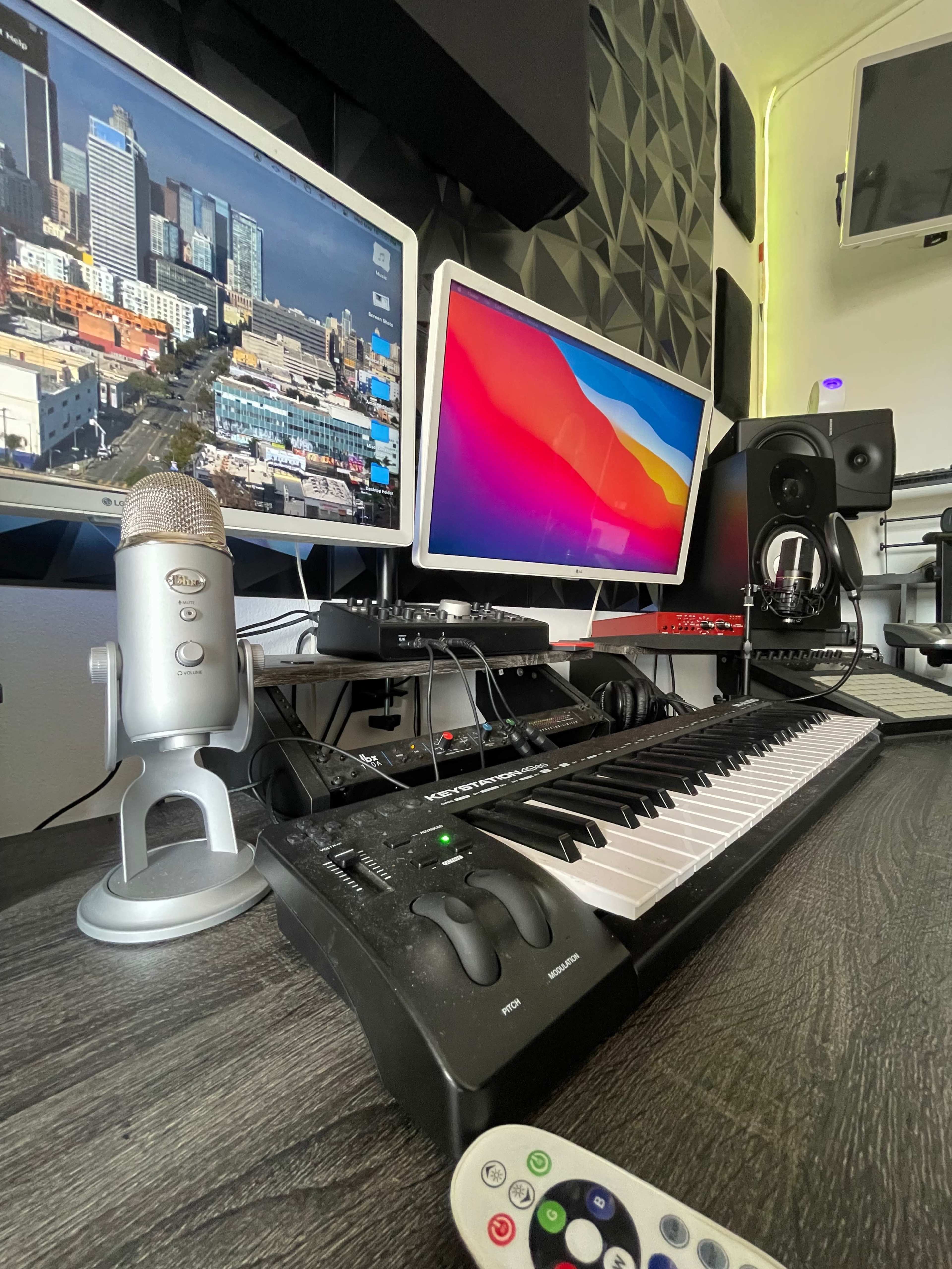 A music production setup features a MIDI keyboard, two computer monitors displaying cityscapes and software, a microphone, and audio equipment on a wooden desk.