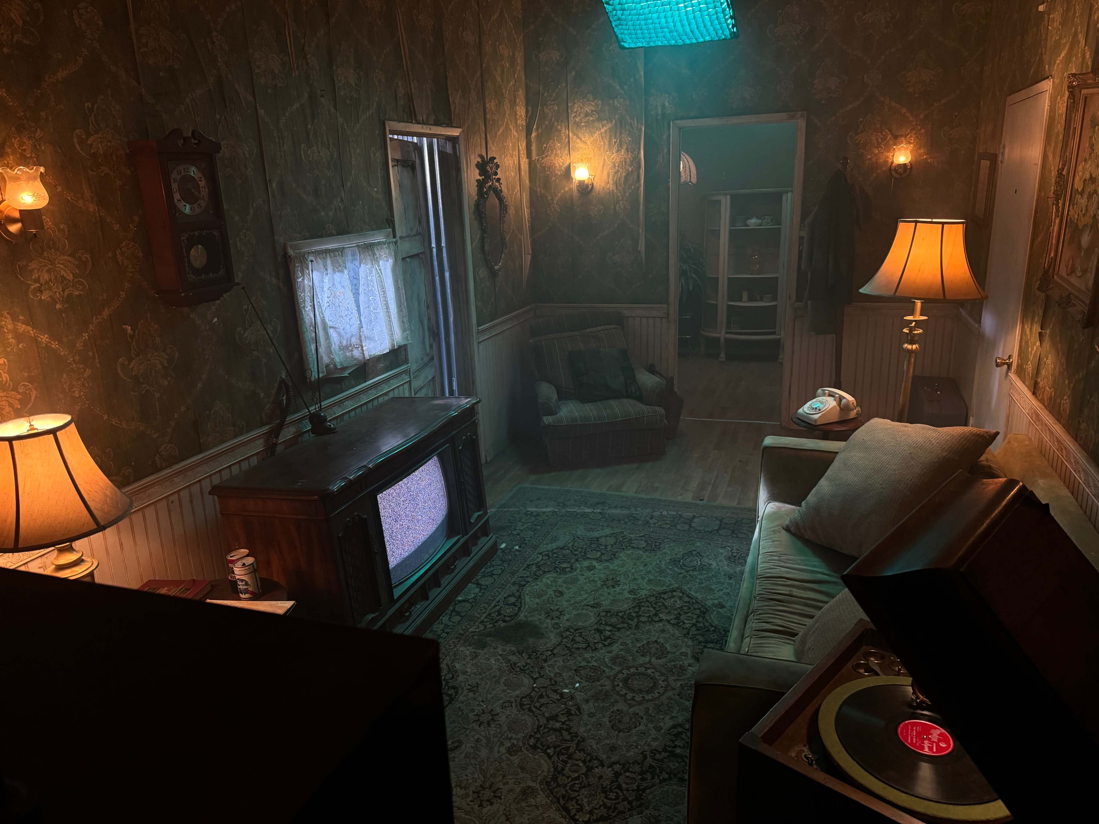 A dimly lit room with vintage furniture, including a sofa, a record player, and a television, all set against patterned wallpaper.
