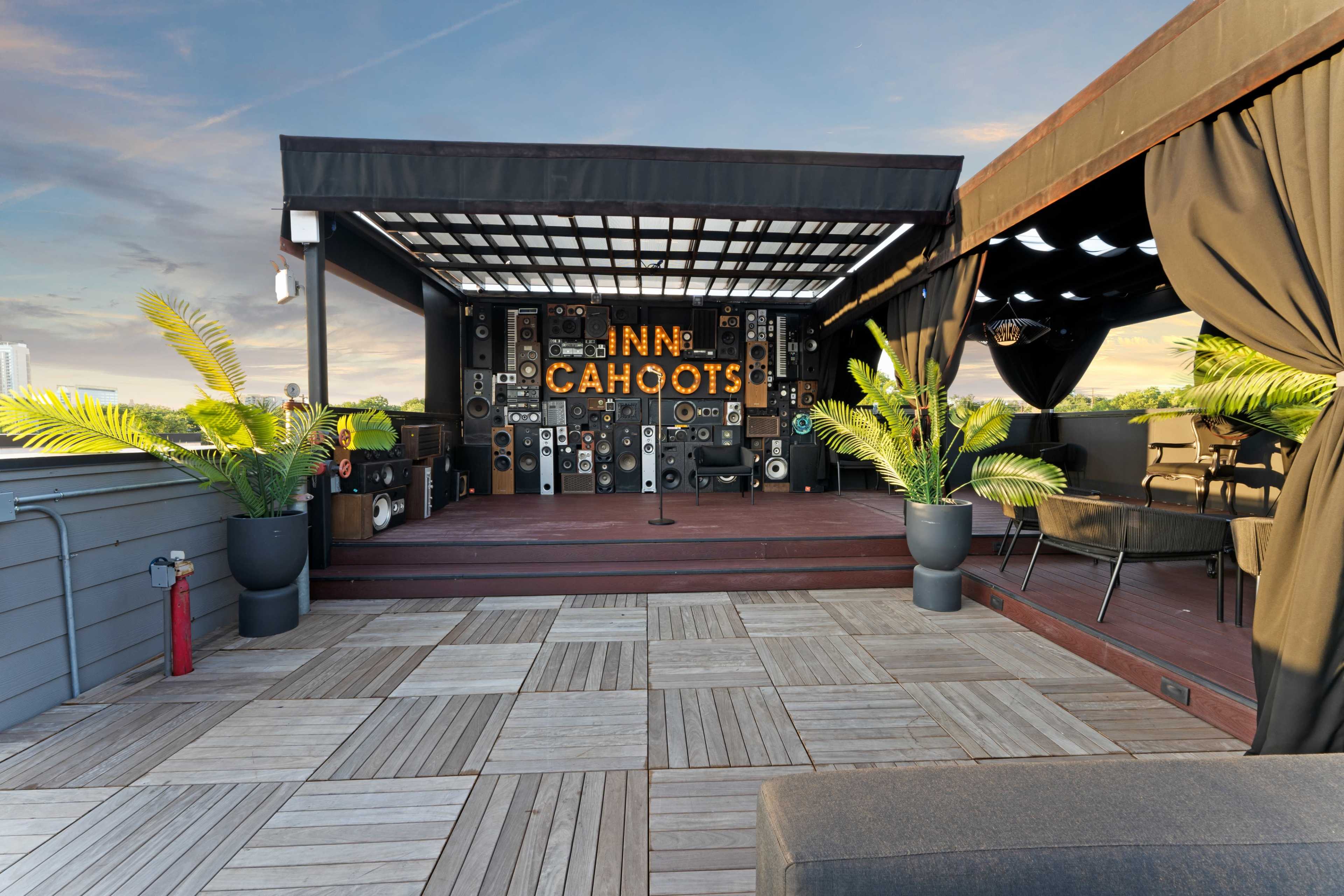 The image shows a rooftop space designed with a stage area featuring a backdrop of large speakers and the word "CAHOOTS," surrounded by potted plants and outdoor seating.