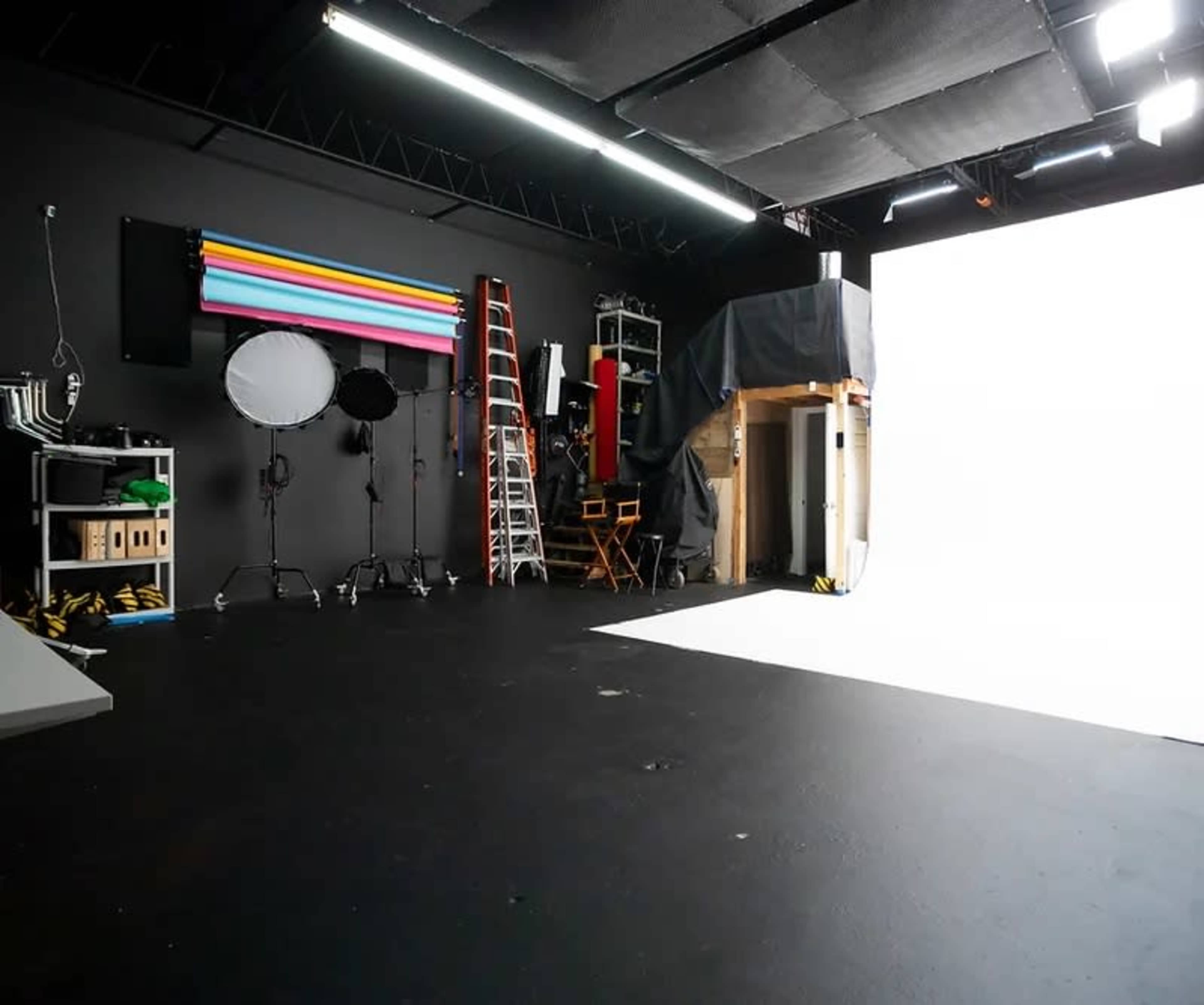 An empty photography studio with various equipment, including lighting, backdrops, and a ladder, set against a white backdrop.