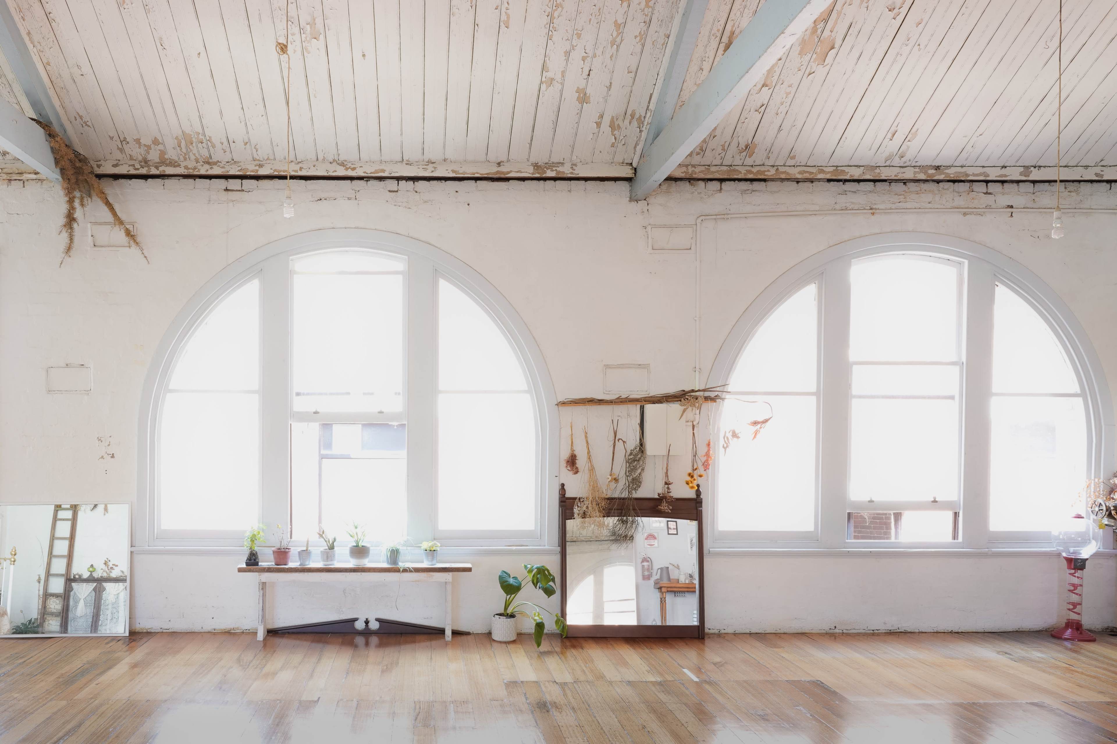 A well-lit room features large arched windows, a reflective surface along the wall, and a simple wooden shelf with potted plants.