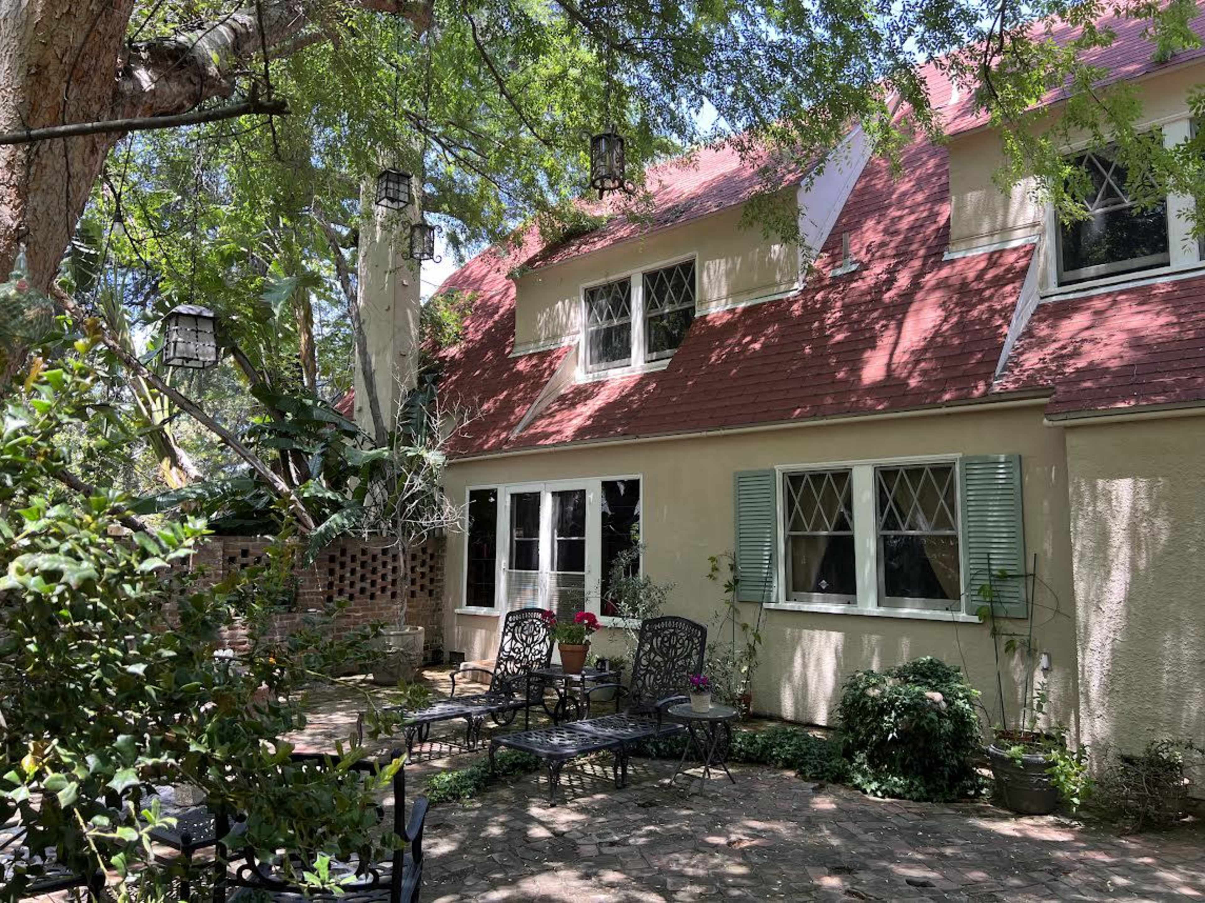 A cozy house with a red sloped roof, surrounded by greenery and patio furniture on a brick floor.