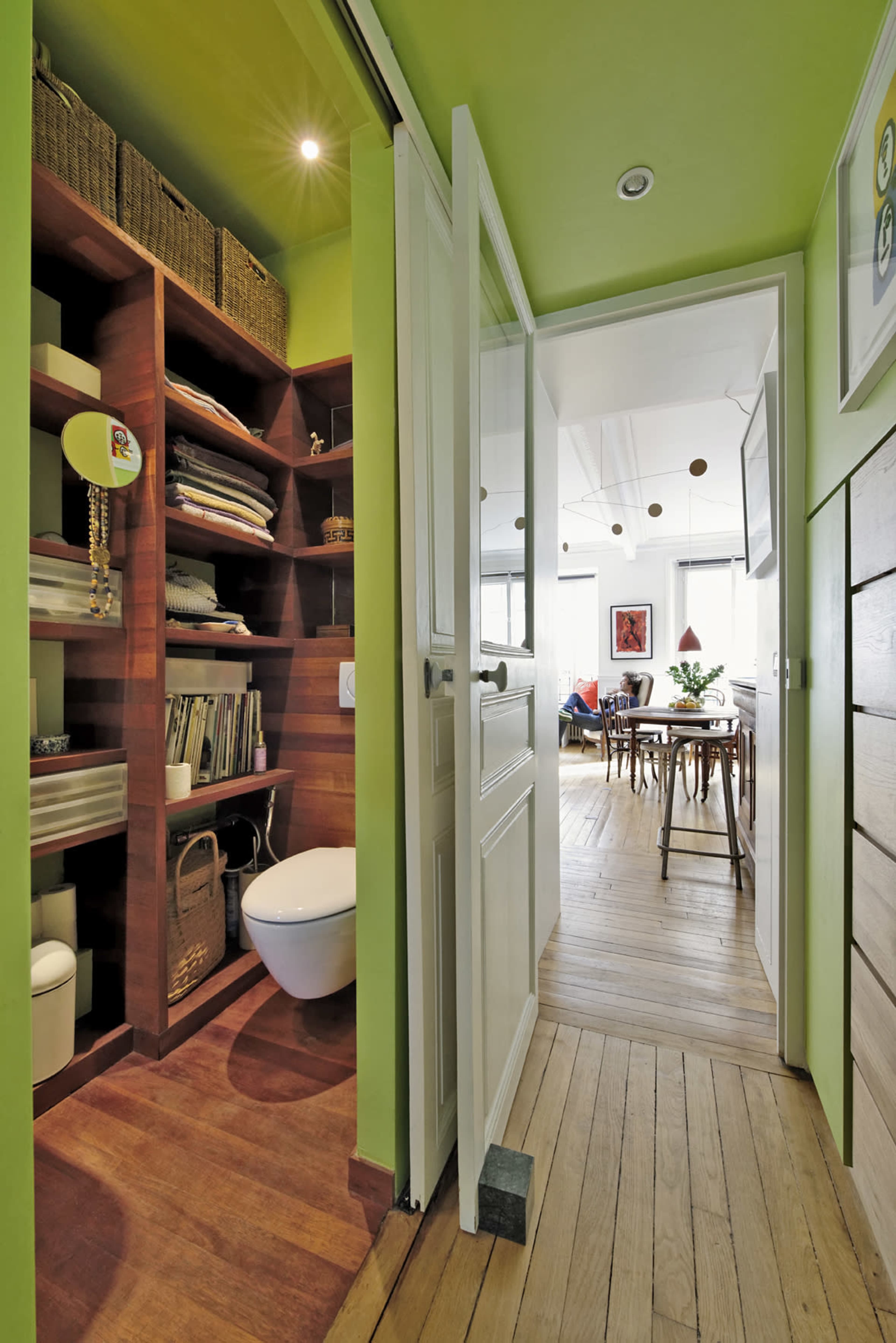 A narrow hallway leads to a bright green bathroom with shelves for storage and an open door that reveals a dining area in the background.