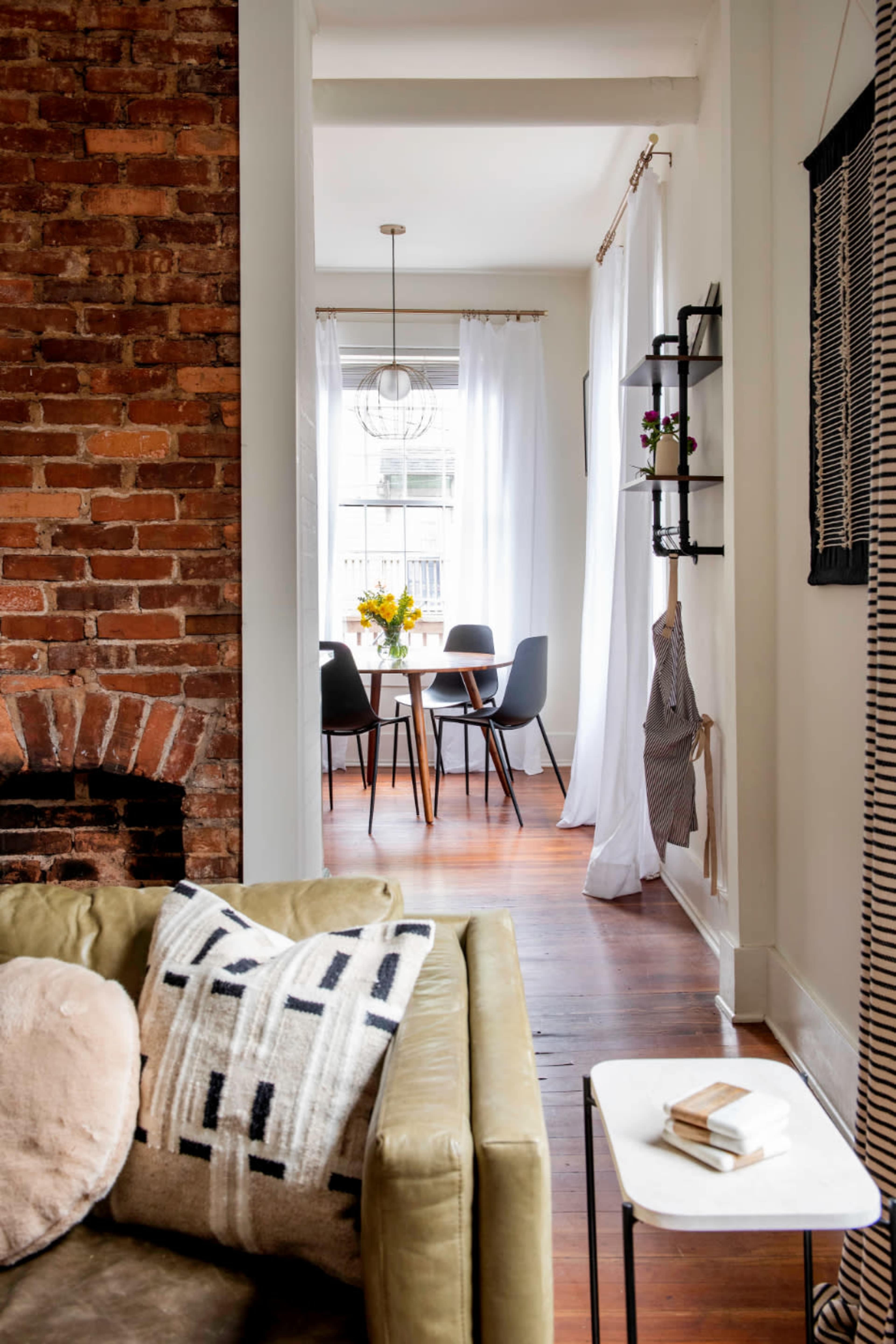 A modern living room features a brick wall and a view of a dining area with a table and chairs beyond sheer curtains.