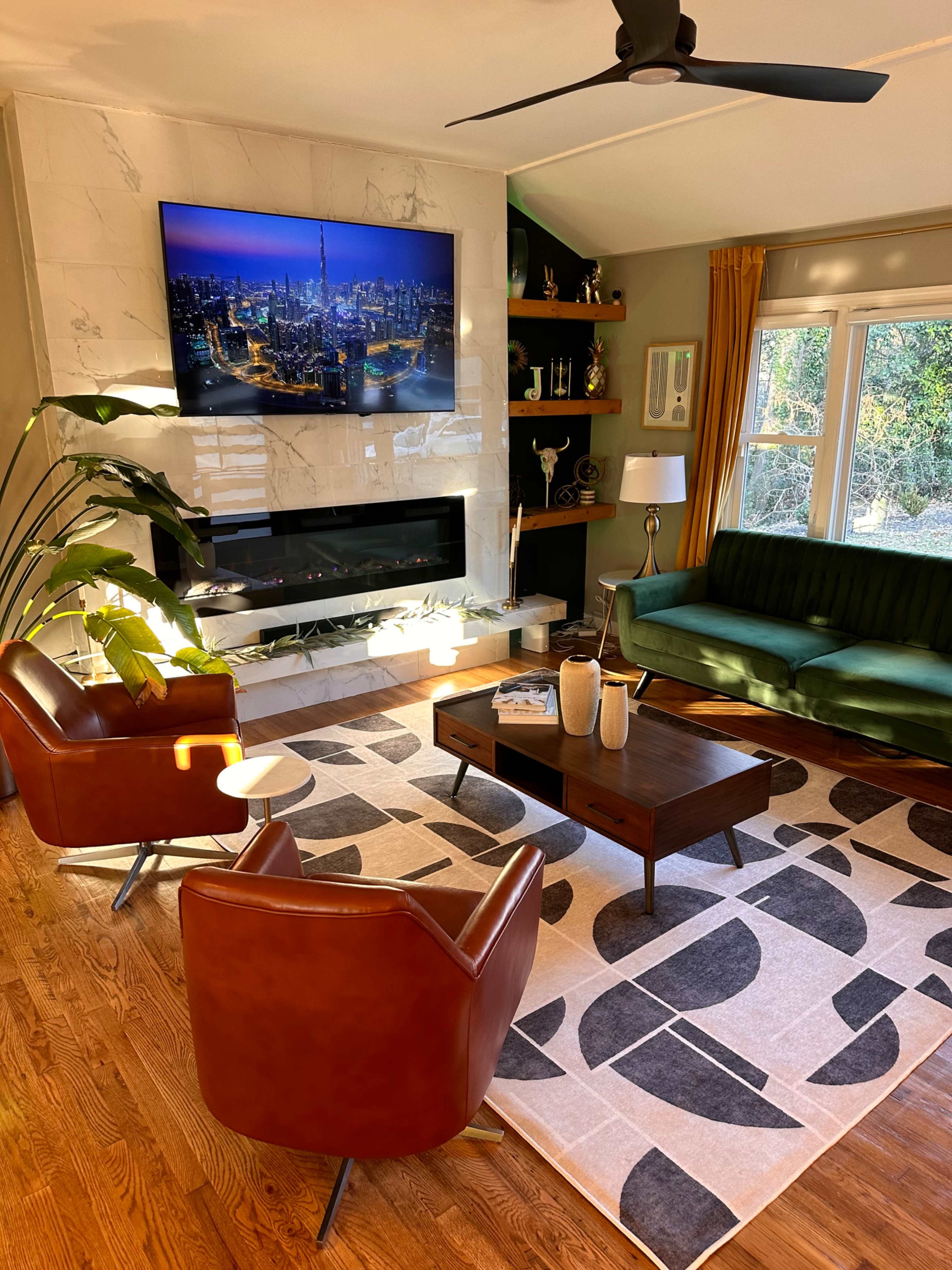 A modern living room featuring a green sofa, two brown armchairs, a wooden coffee table, and a large television mounted above a marble fireplace.