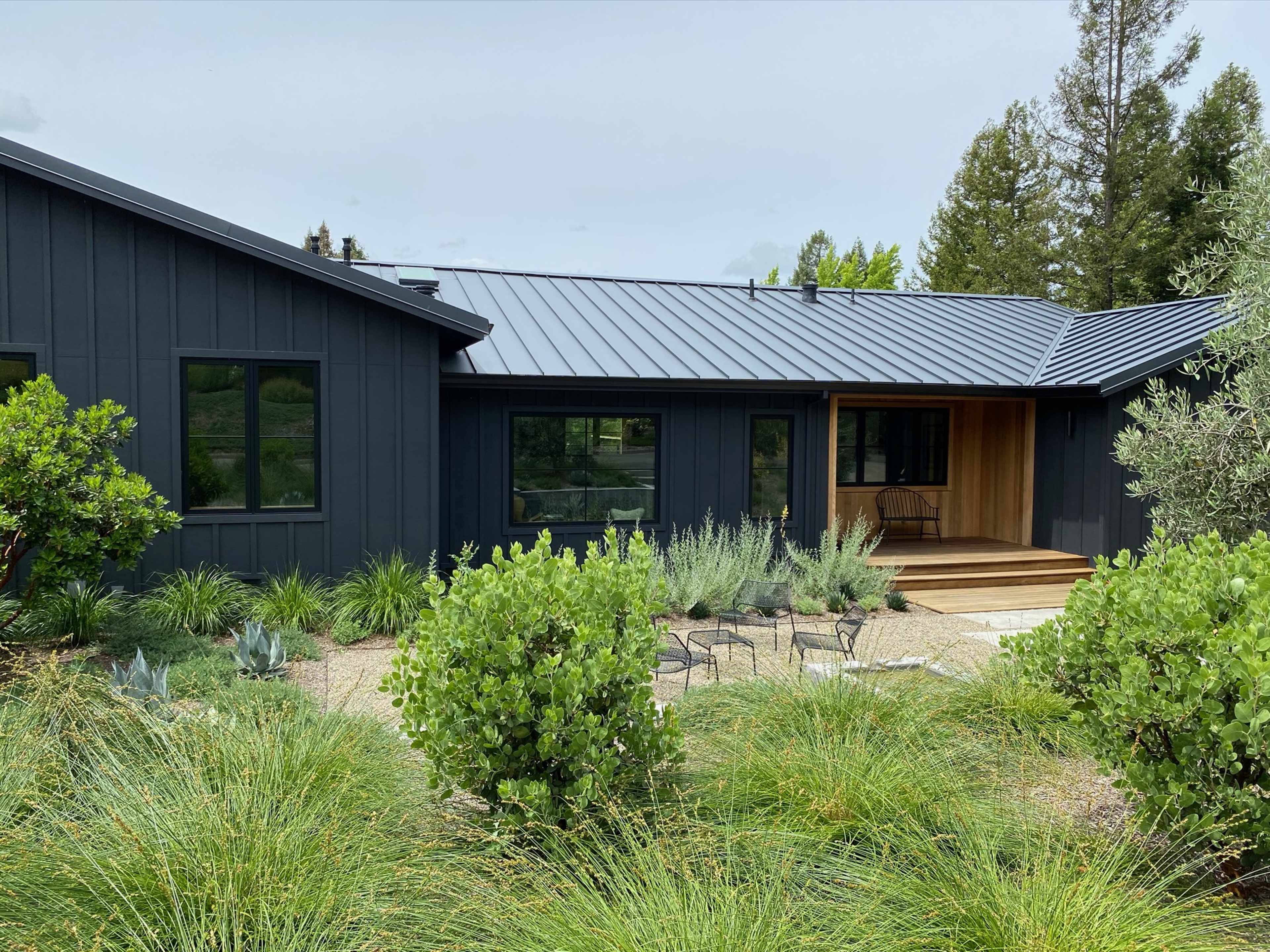 A modern black-clad house features a wooden front porch, surrounded by landscaped greenery and decorative plants.
