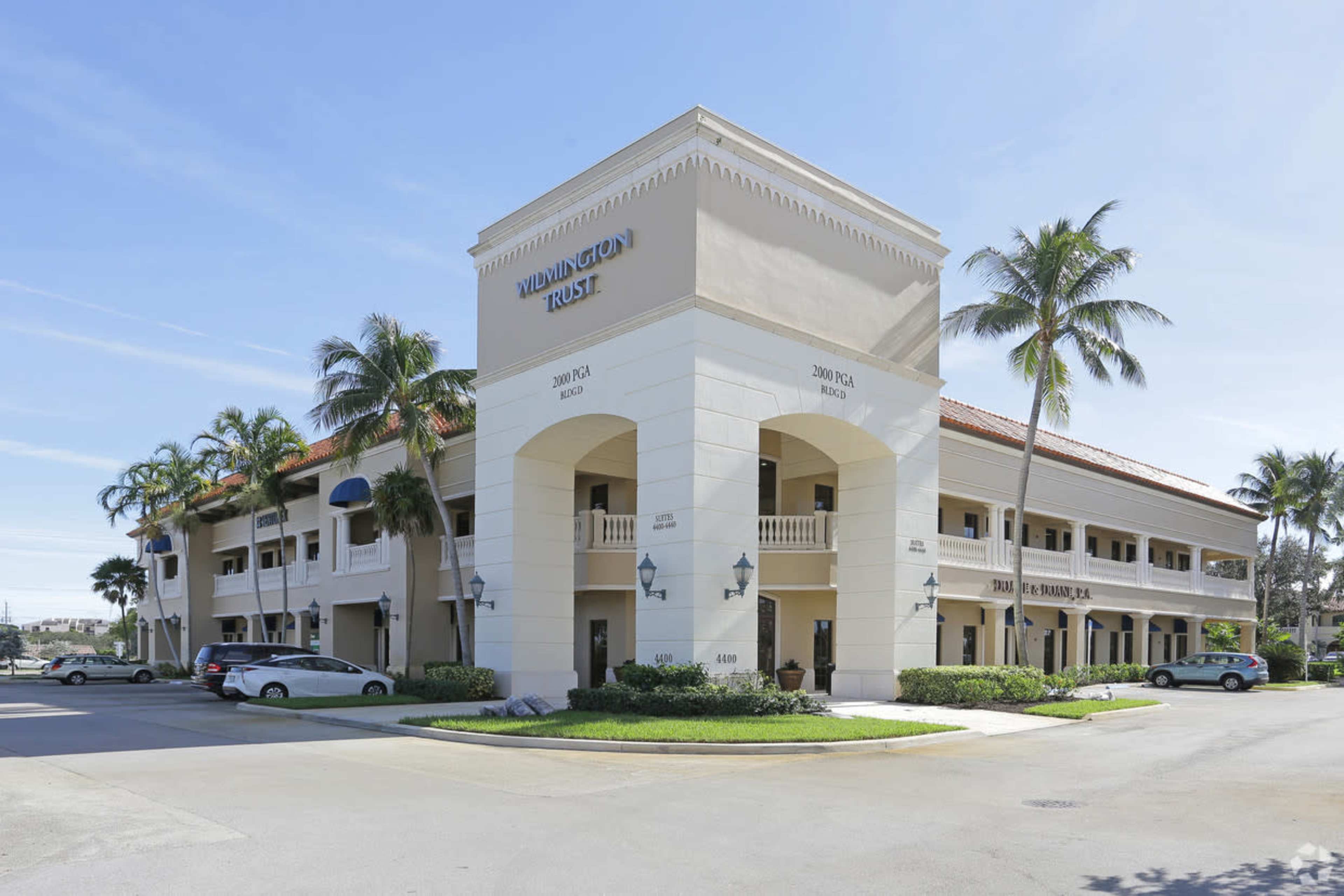 The image shows a two-story office building with a light-colored facade, palm trees, and cars parked in front.