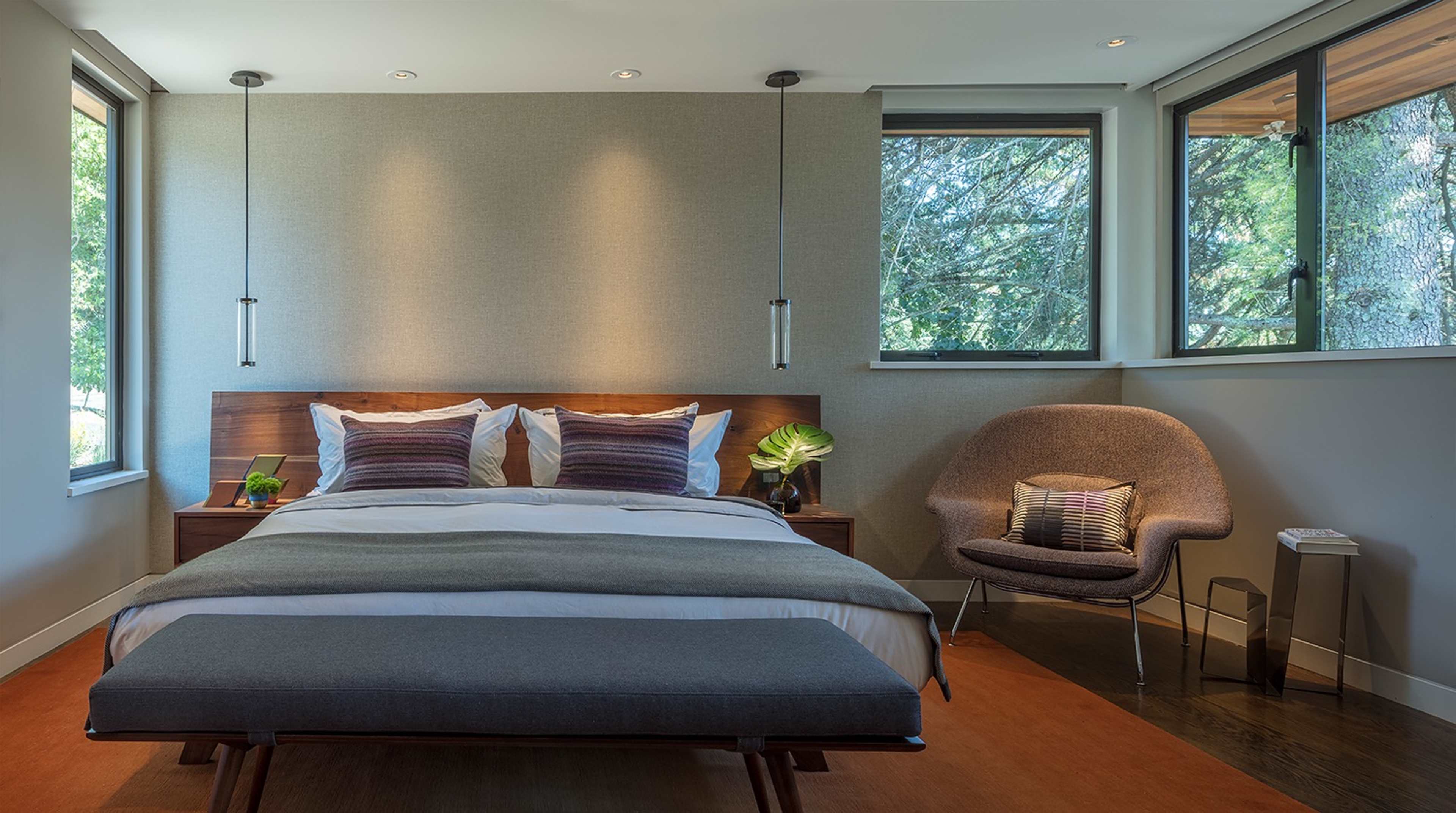 A modern bedroom features a wooden bed with white linens, a gray bench at the foot, and a cozy chair beside large windows.
