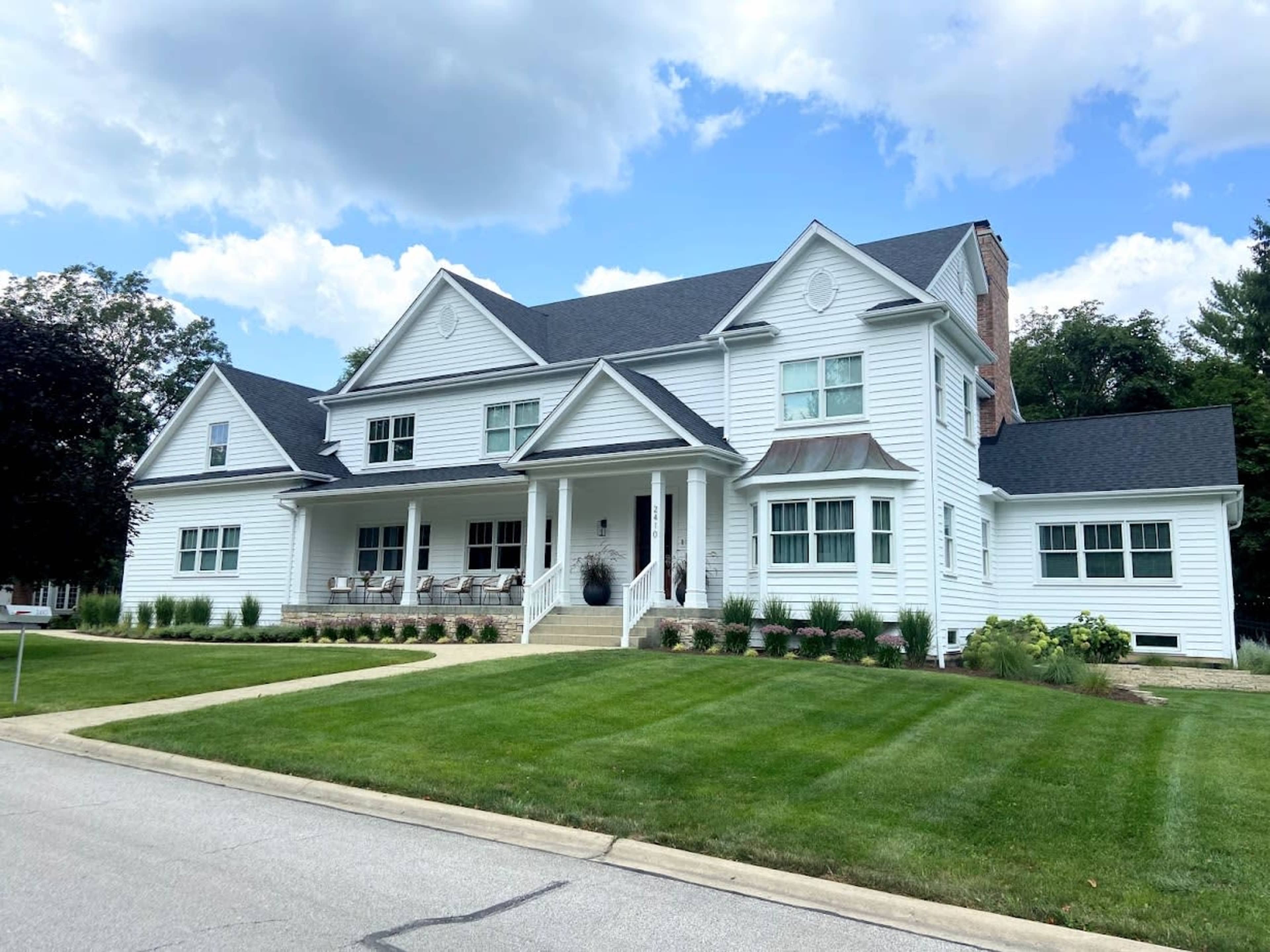 The image shows a large, two-story white house with a gray roof, featuring a front porch and well-maintained lawn.