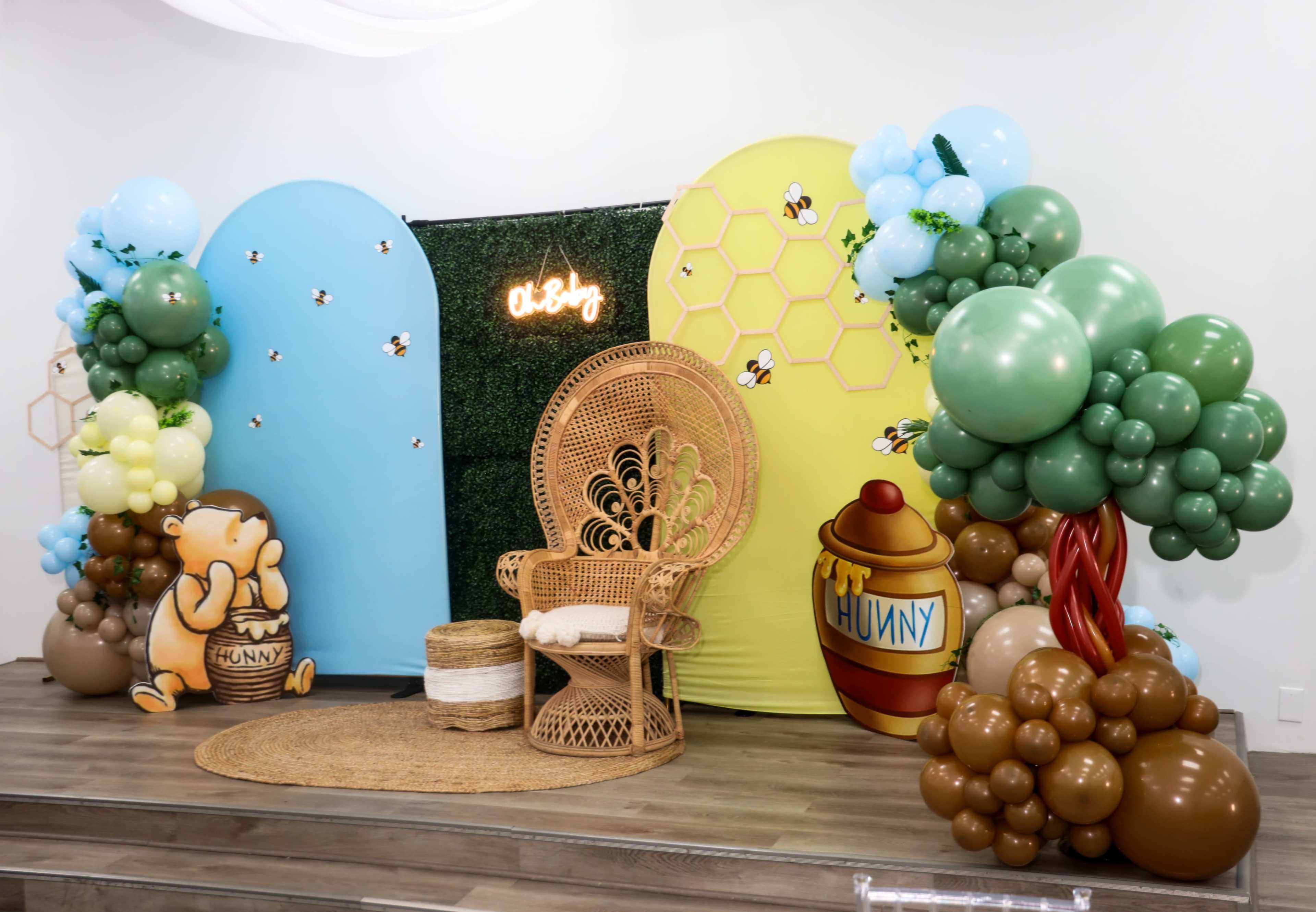 The image shows a themed party backdrop featuring colorful decorations, a wicker chair, and balloon arrangements resembling trees and honey elements.