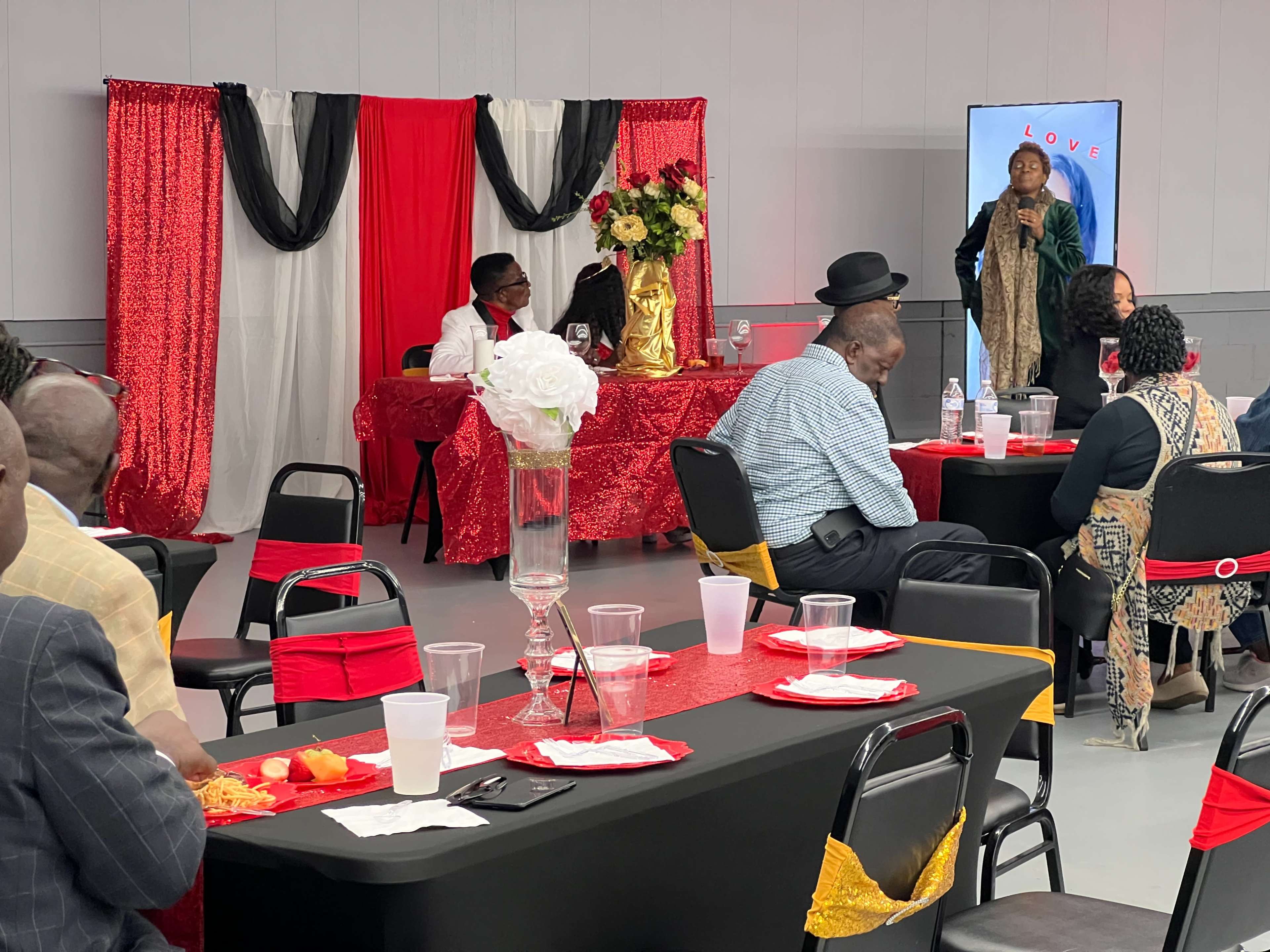 A group of people is seated at tables with red and black tablecloths while a speaker addresses the audience in a decorated room.