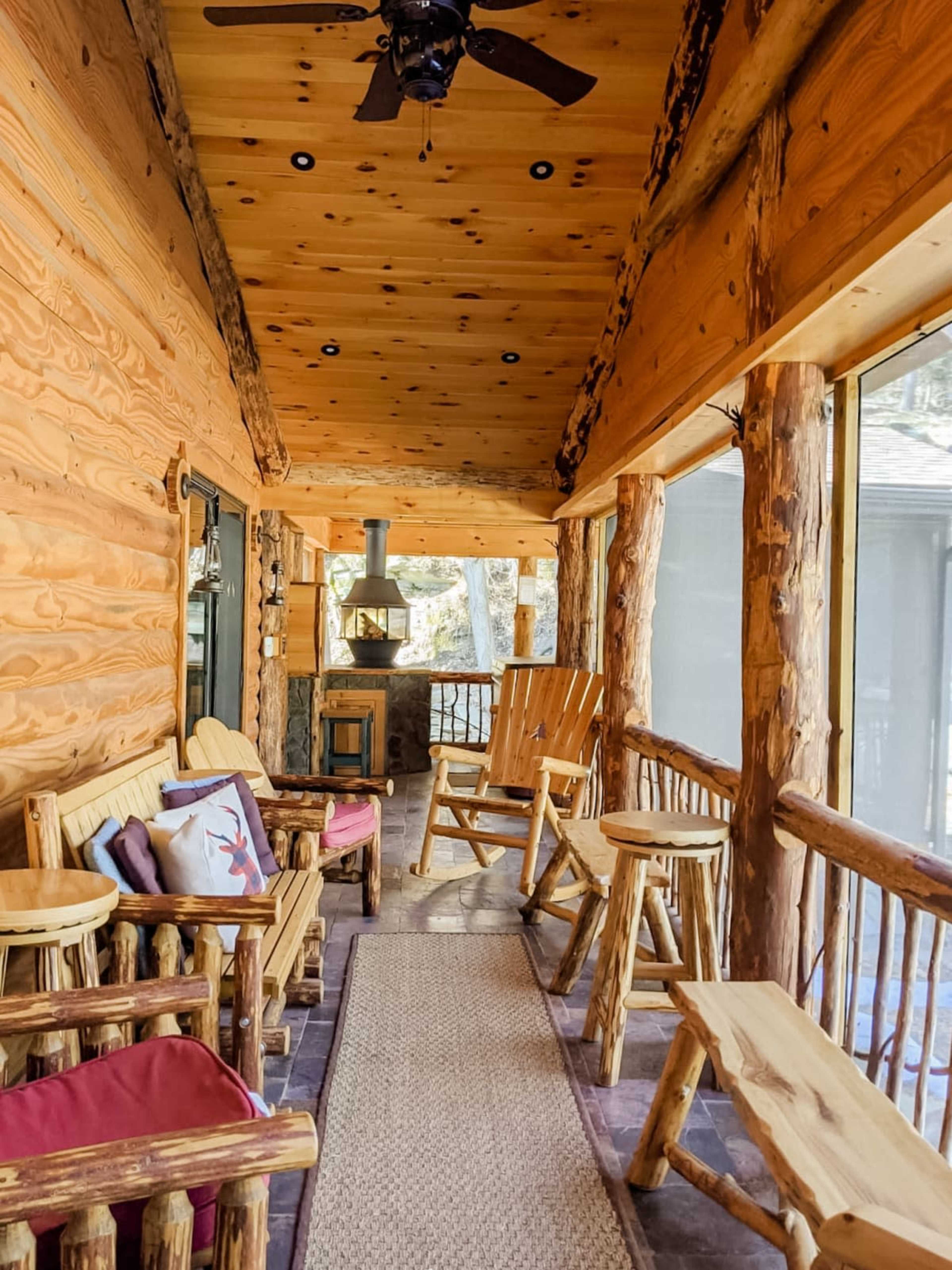 A wooden porch features log furniture, a ceiling fan, and a cozy seating area surrounded by windows.