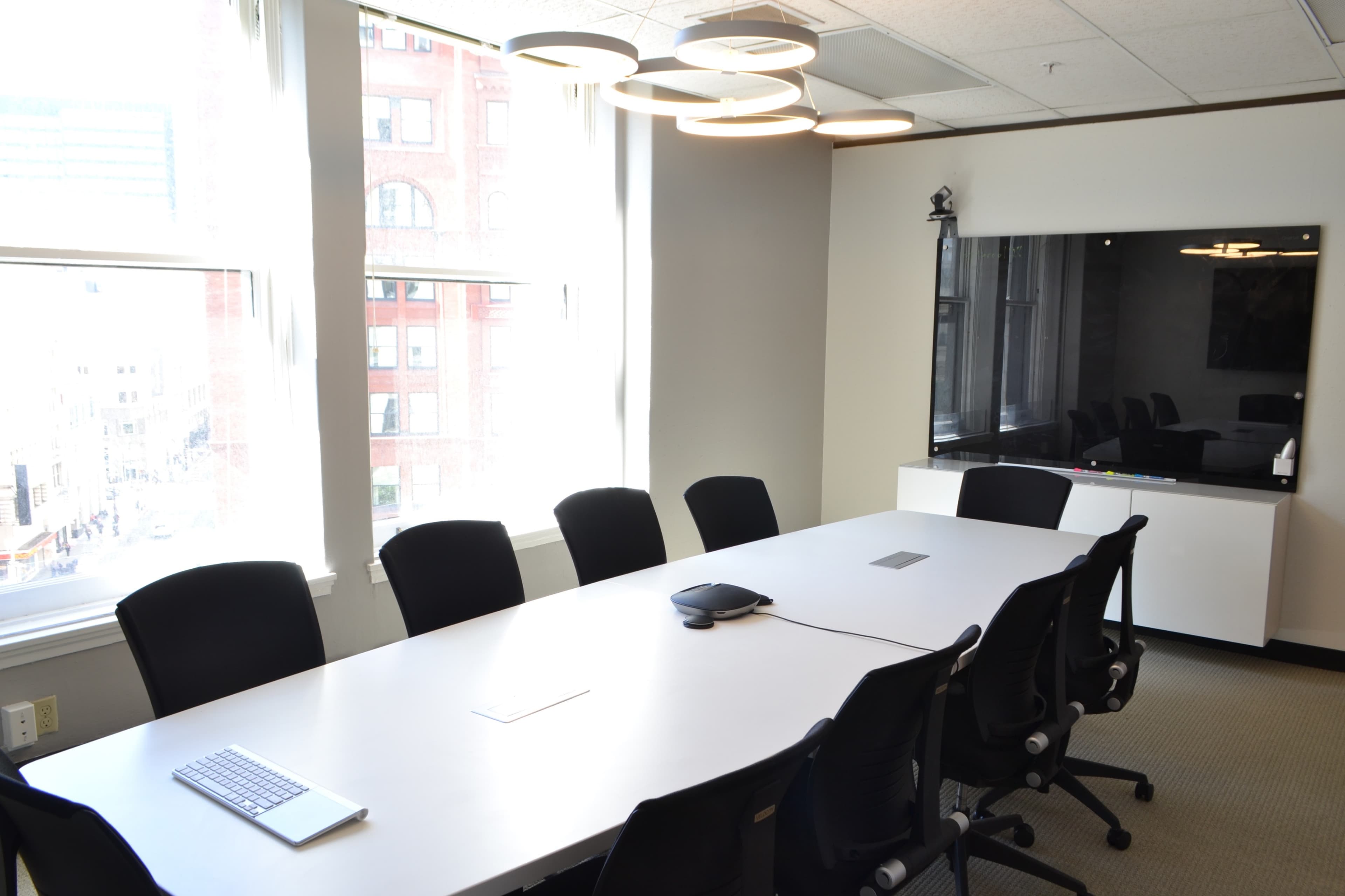 A conference room features a long white table surrounded by black chairs, with large windows allowing natural light and a black screen mounted on the wall.