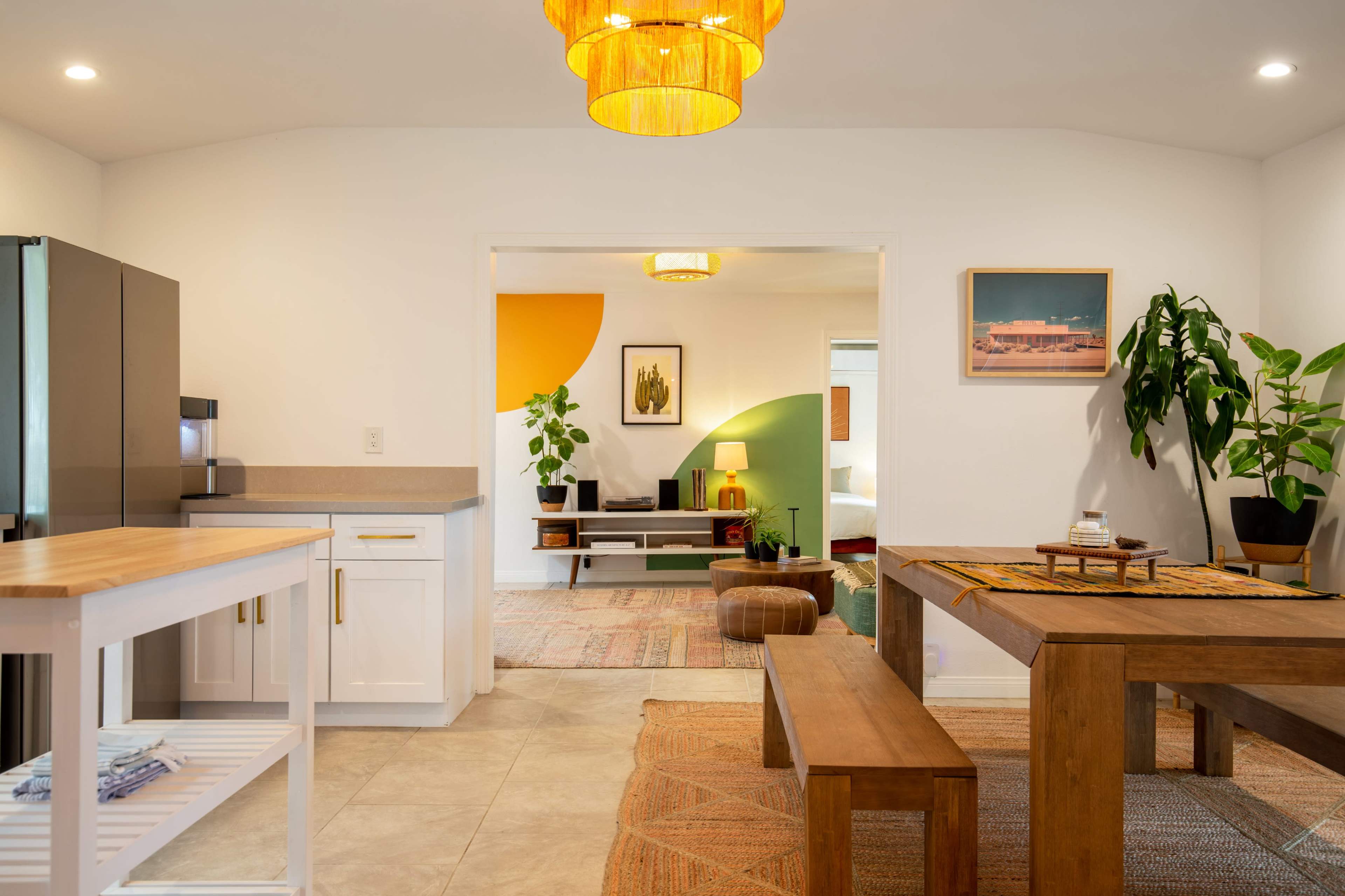 The image features a modern kitchen and dining area, highlighted by a wooden dining table, a refrigerator, and decorative plants in a brightly lit space.