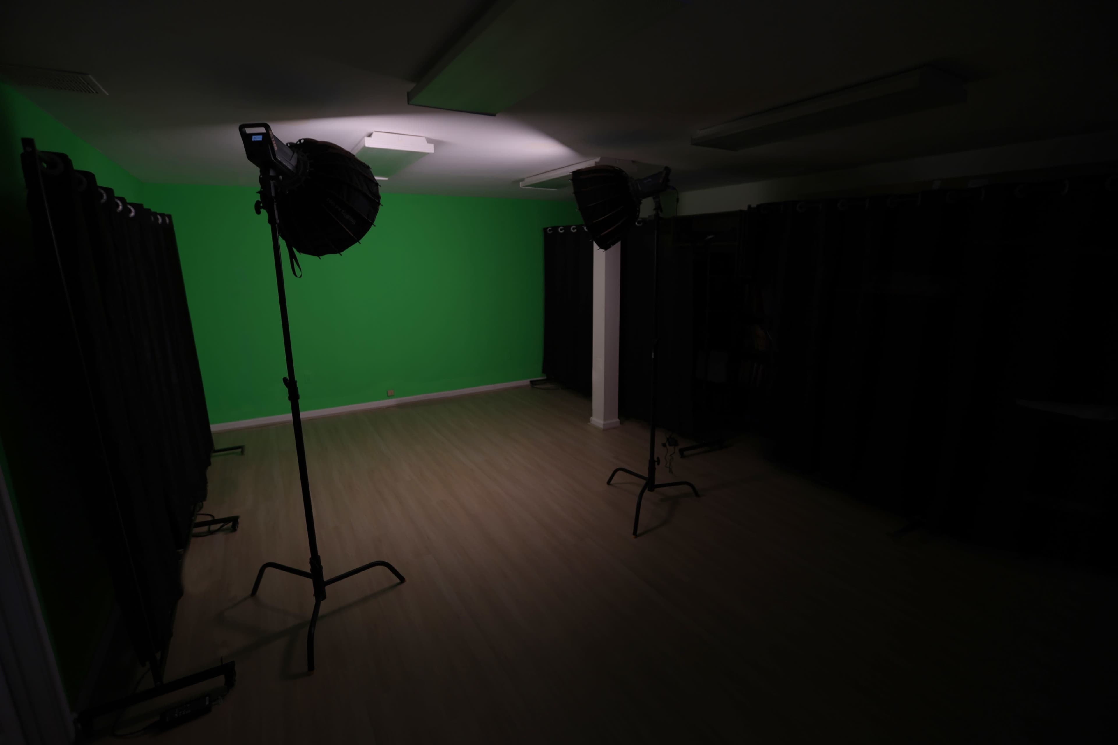 The image shows an empty studio space with a green screen on one wall and two lighting setups positioned on stands.