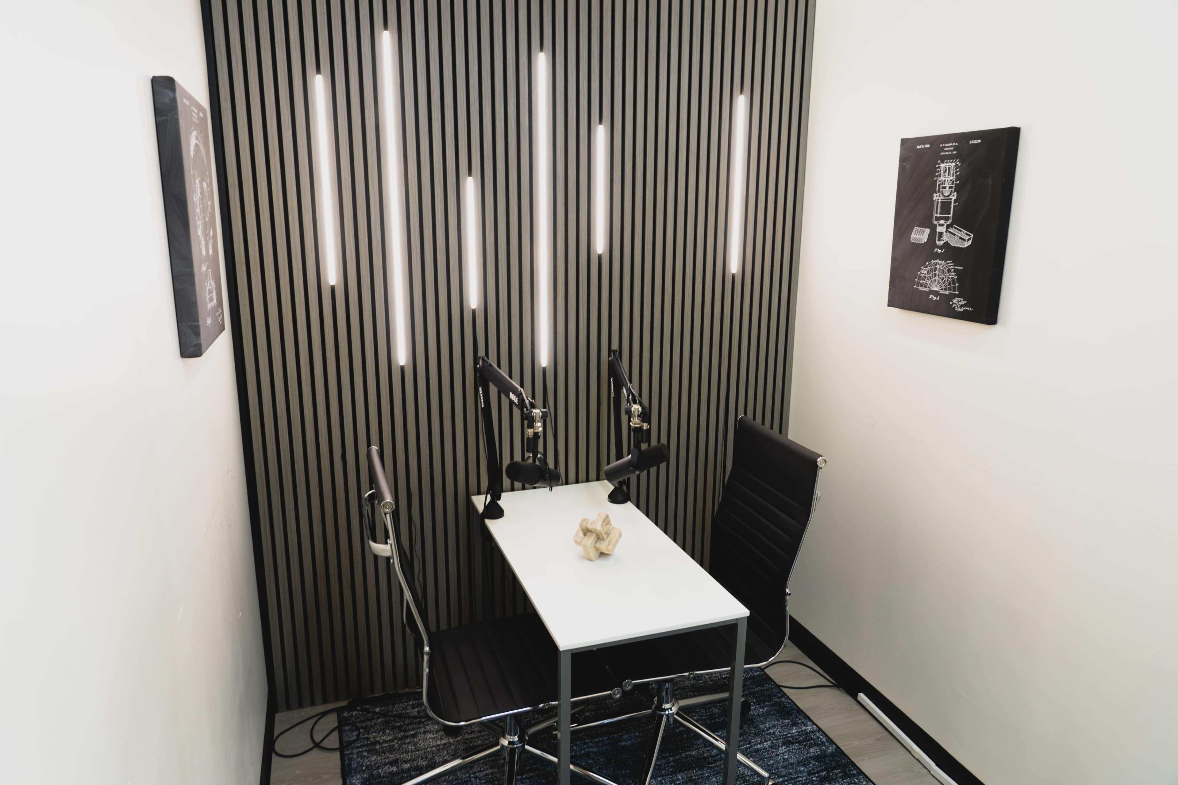 The image shows a small podcast recording studio featuring a white table, two black chairs, and wall-mounted microphones, illuminated by vertical light strips.