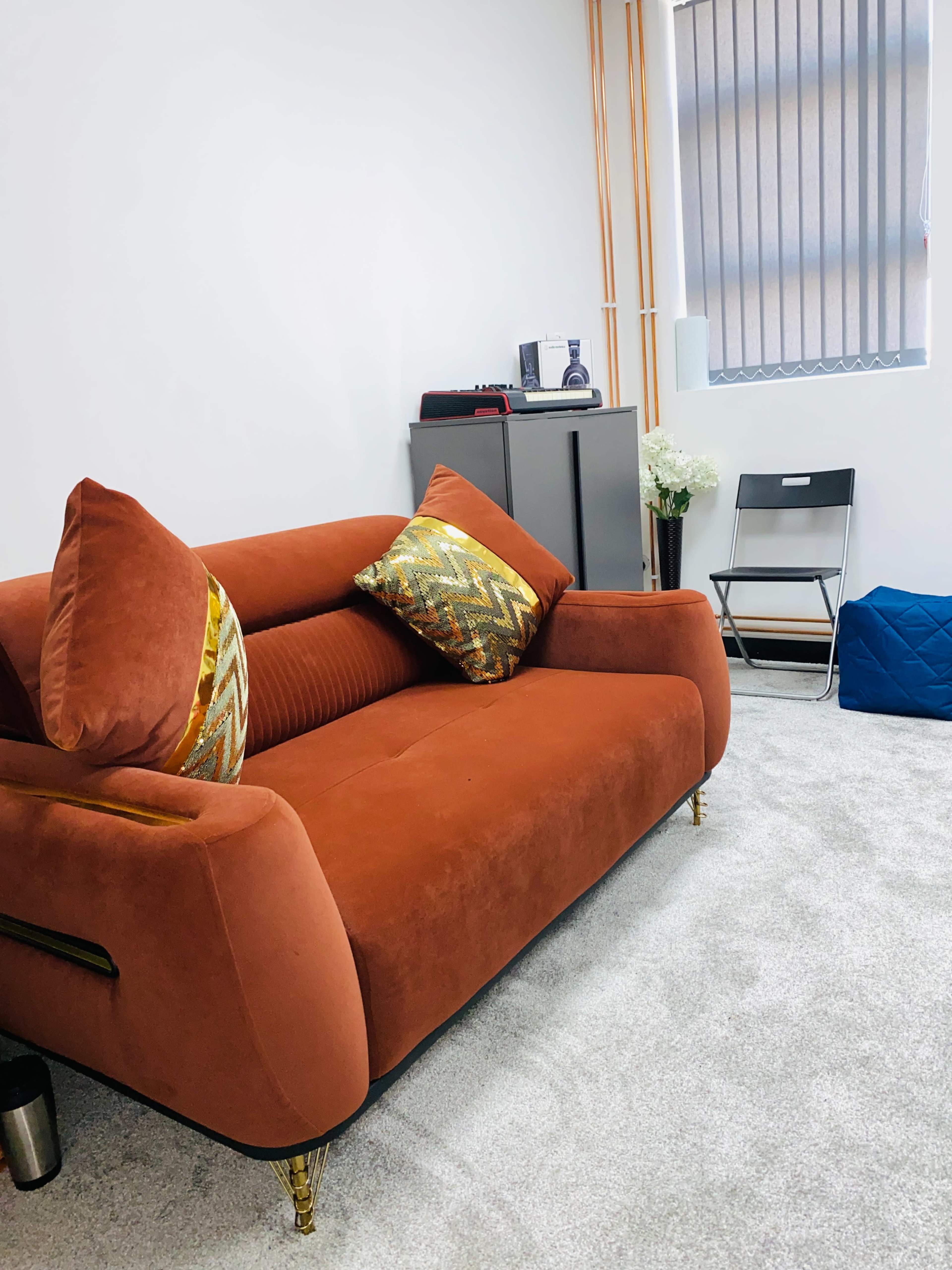 The image shows an orange sofa with decorative pillows positioned in a light-colored carpeted room that includes a chair and a cabinet.