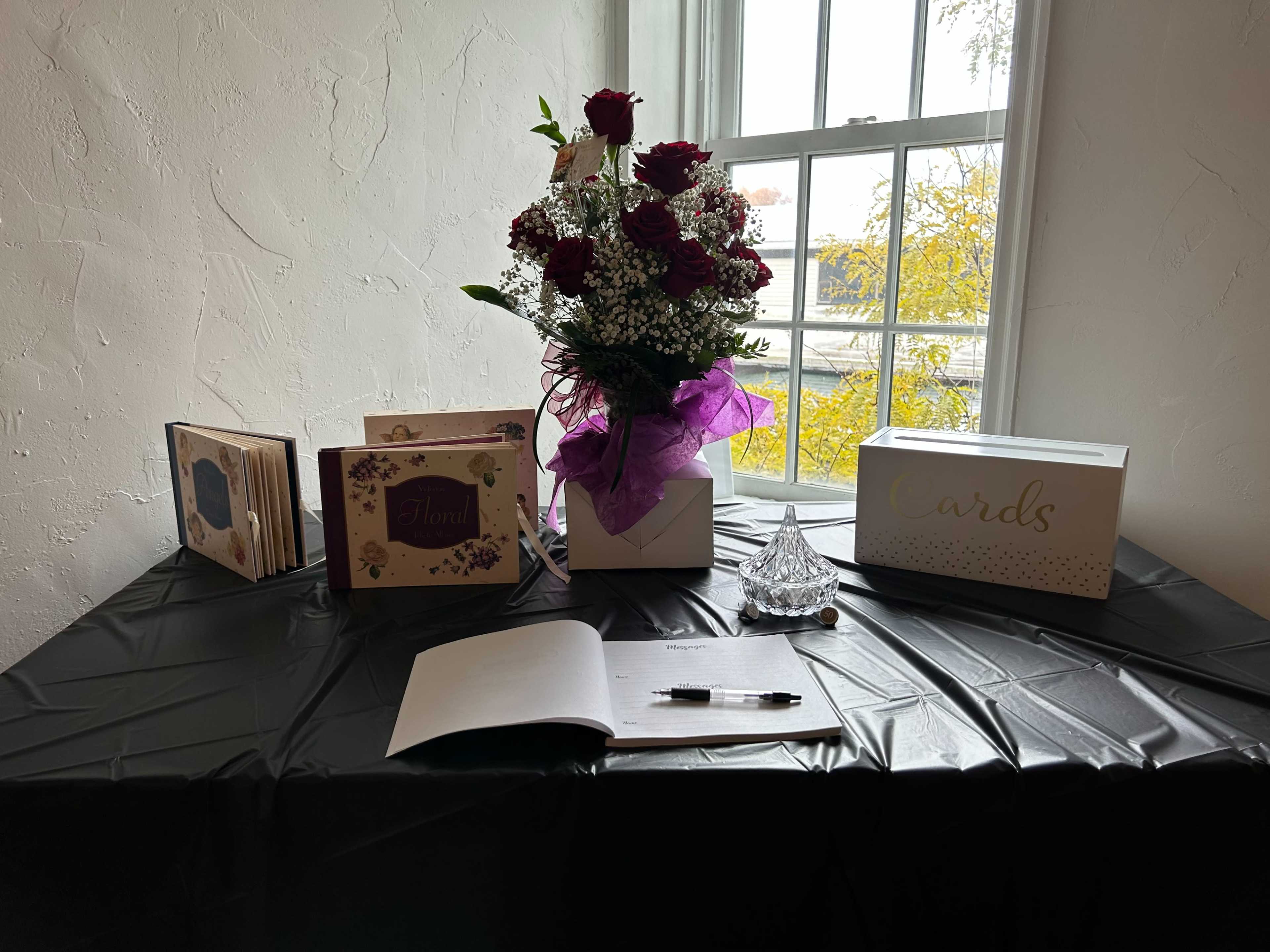 A table with a black tablecloth displays a bouquet of roses, two guest books, a card box, and a pen, all set near a window.