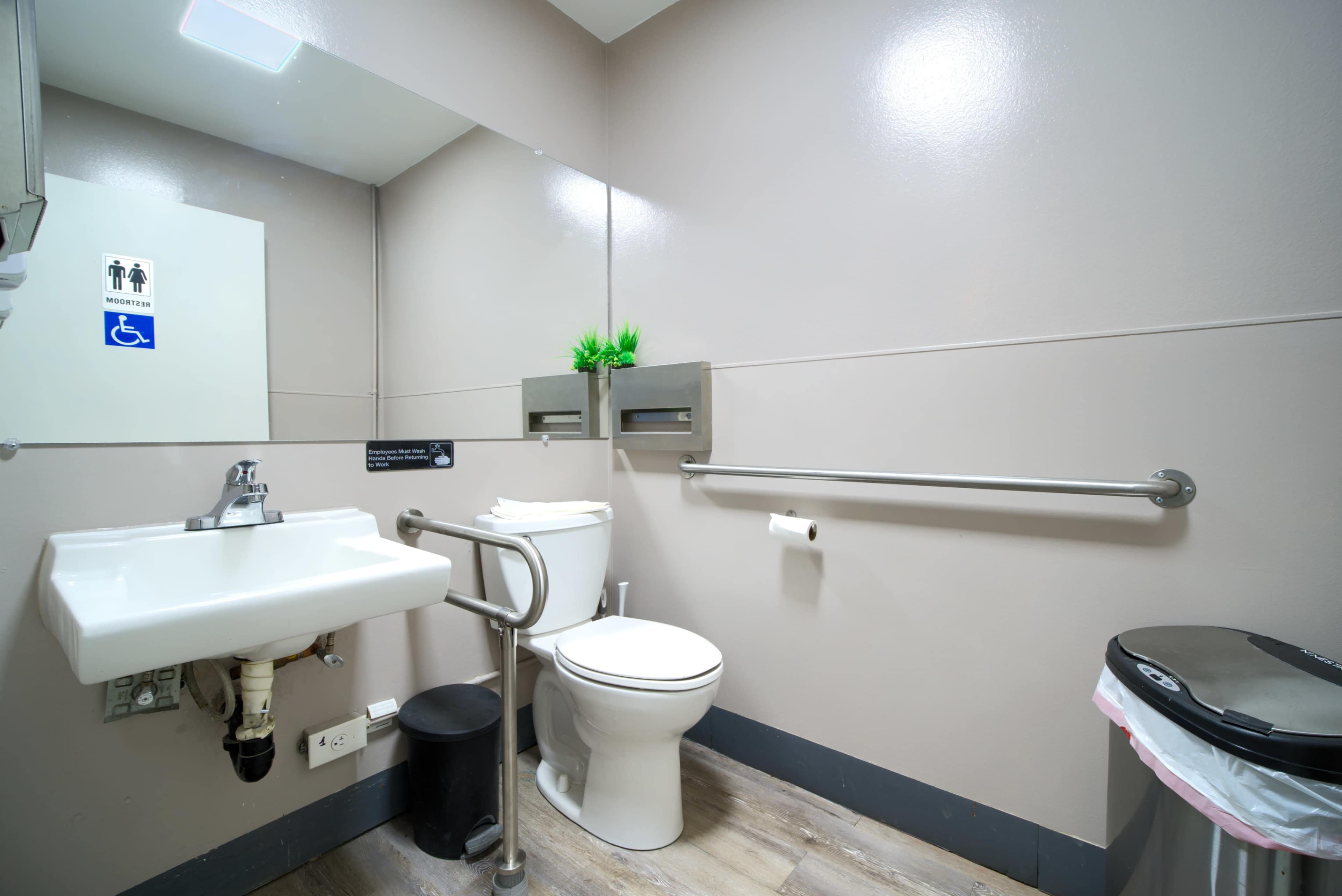 The image shows a clean restroom with a toilet, sink, grab bars, and a trash bin.