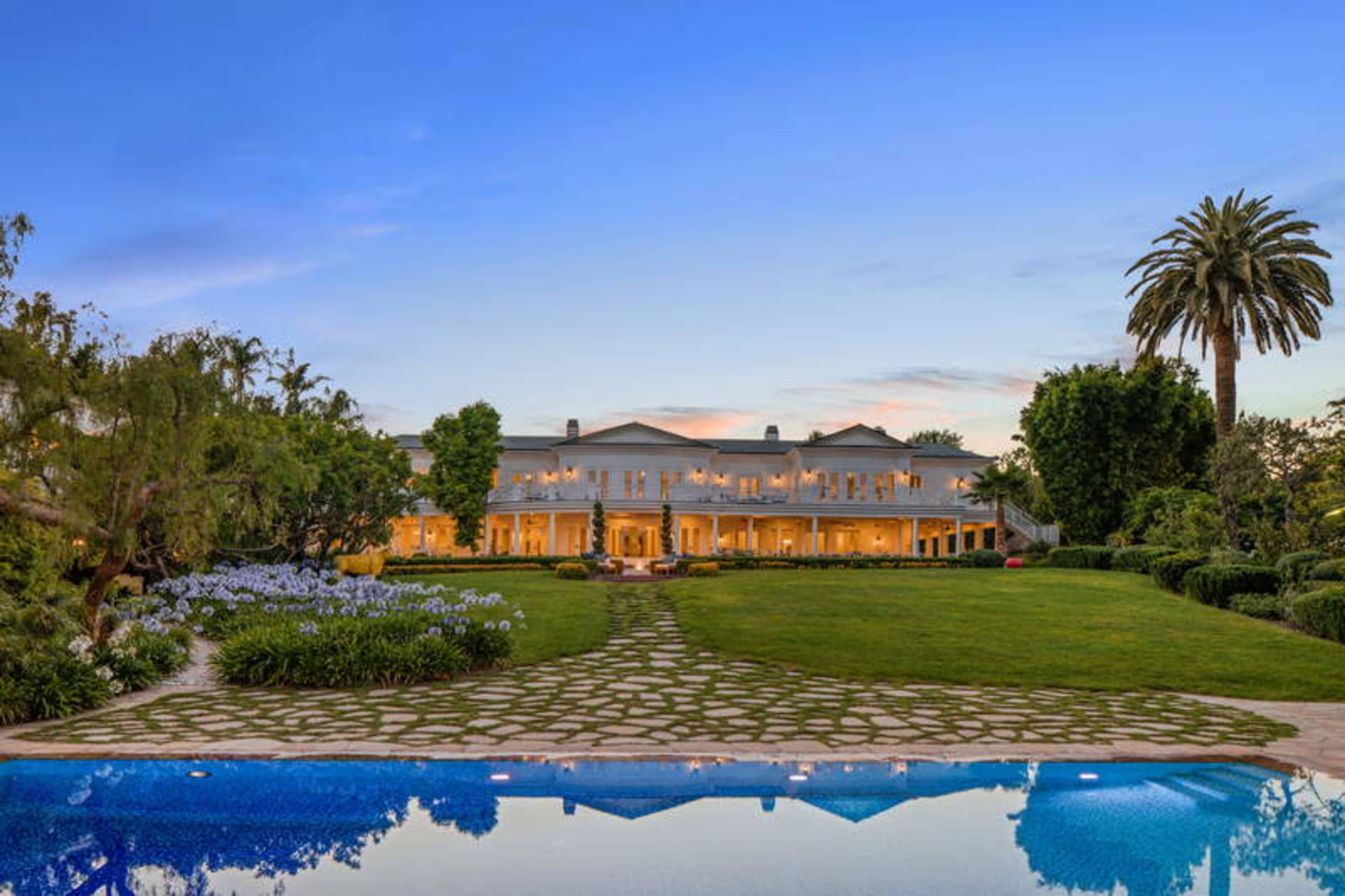 A large, two-story house with a wide porch overlooks a lush lawn and a swimming pool, surrounded by trees and flowers under a clear blue sky.