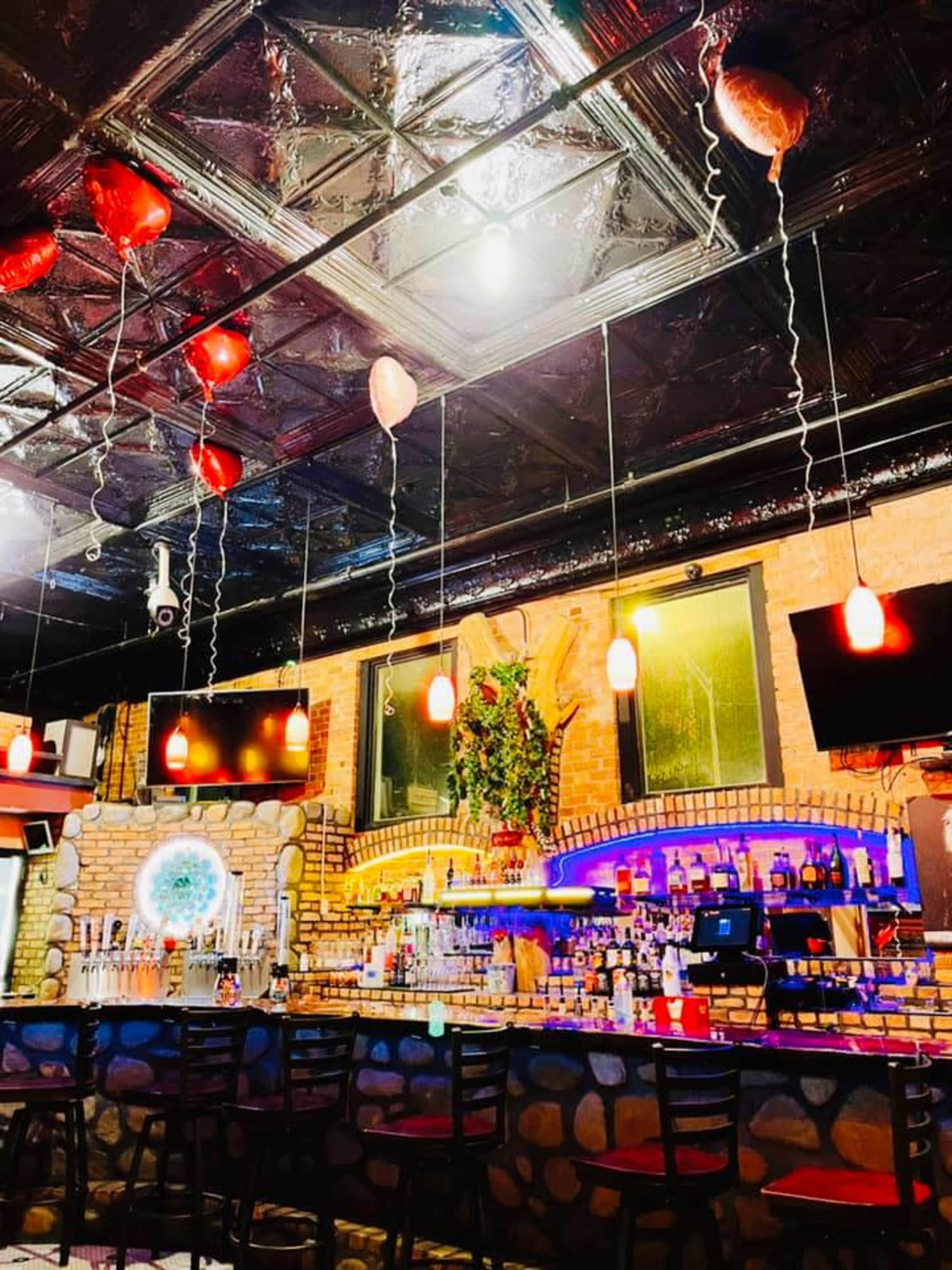 A bar interior with a stone wall, a well-stocked bar counter, and red heart-shaped balloons hanging from the ceiling.