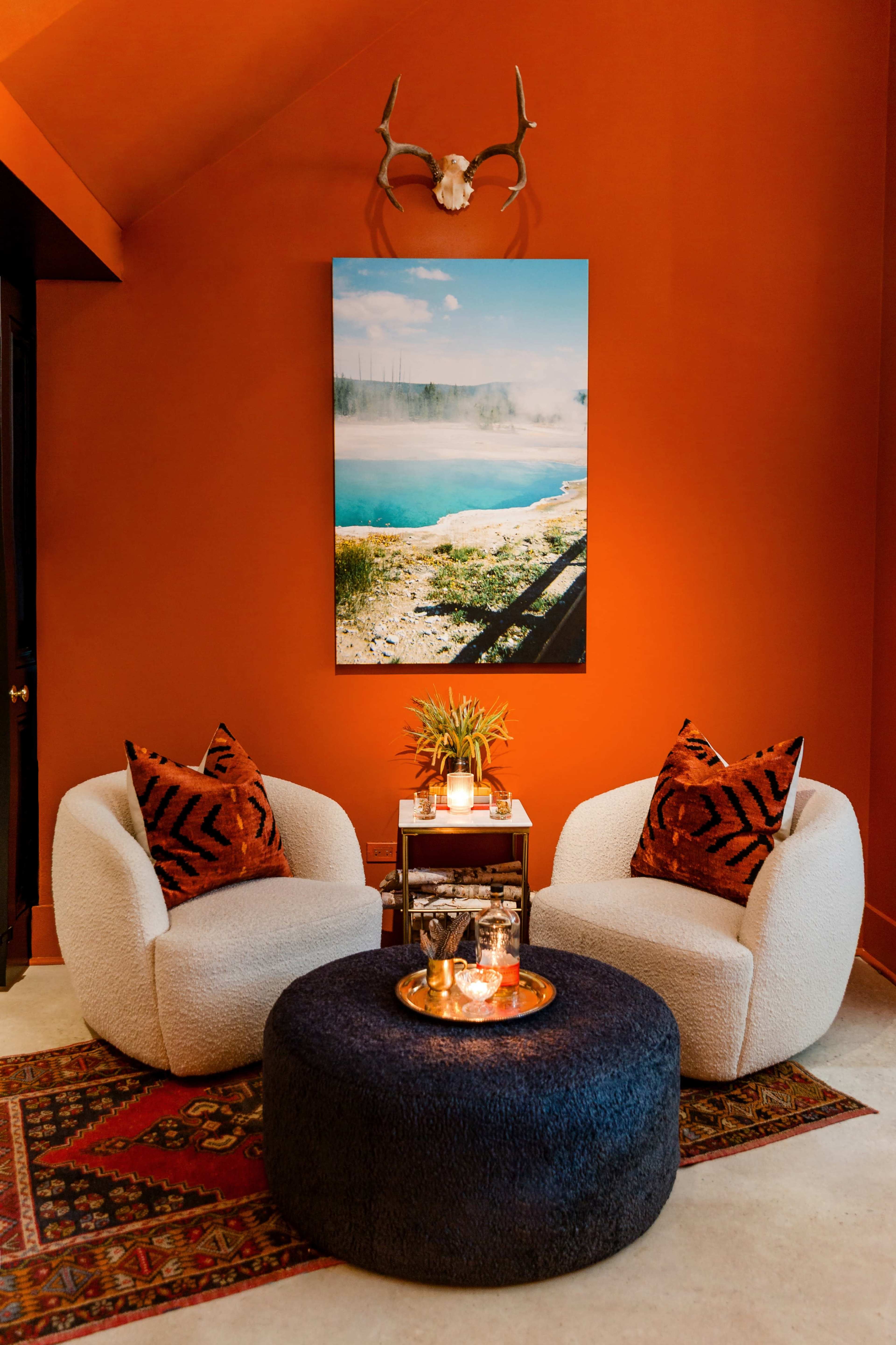 A cozy seating area features two plush chairs with patterned cushions, a round dark ottoman, and a small table adorned with candles and flowers, set against an orange wall with a large landscape photograph above.