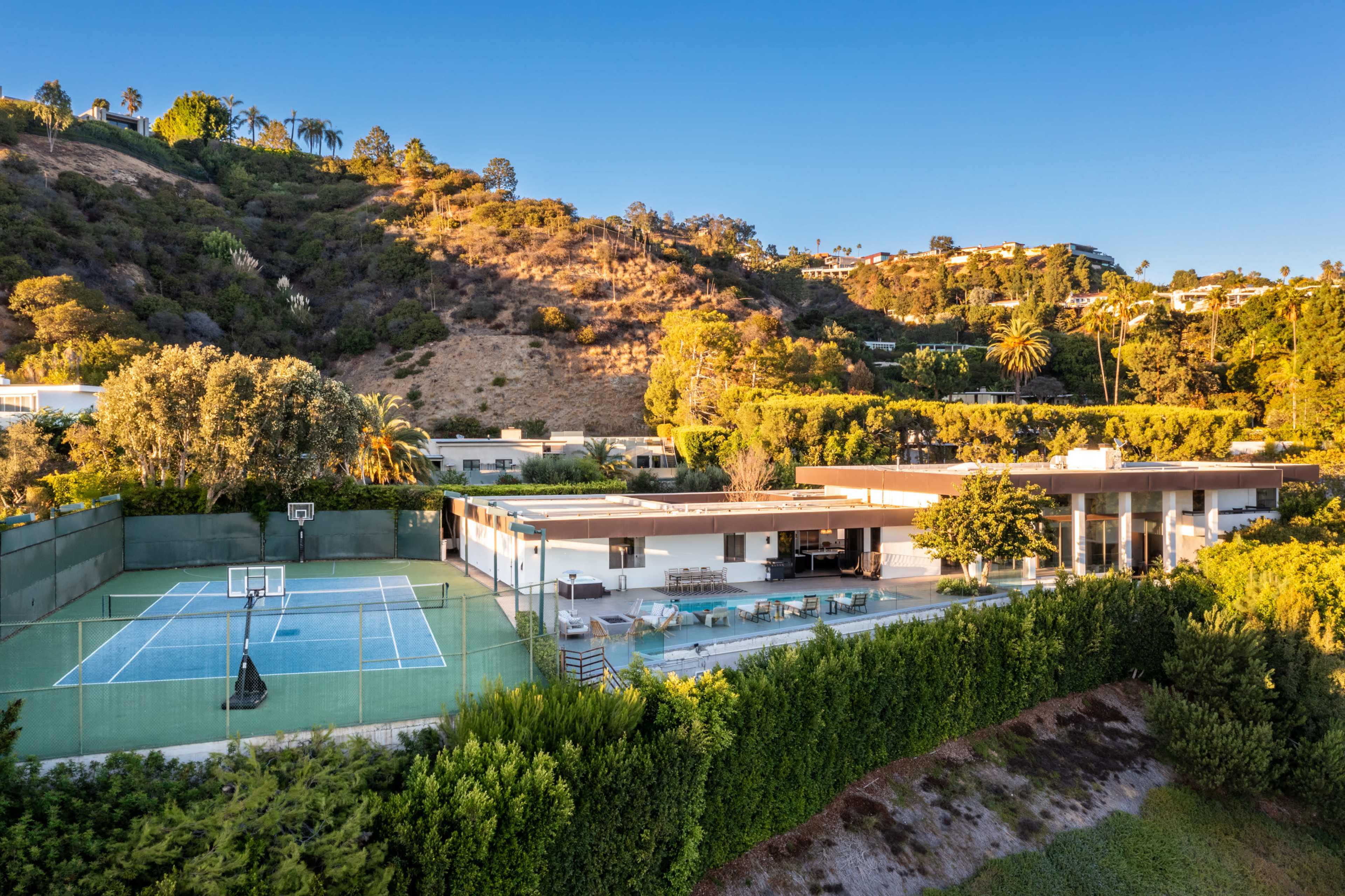 A modern house with a basketball court and tennis court is situated at the base of a hillside covered in greenery.