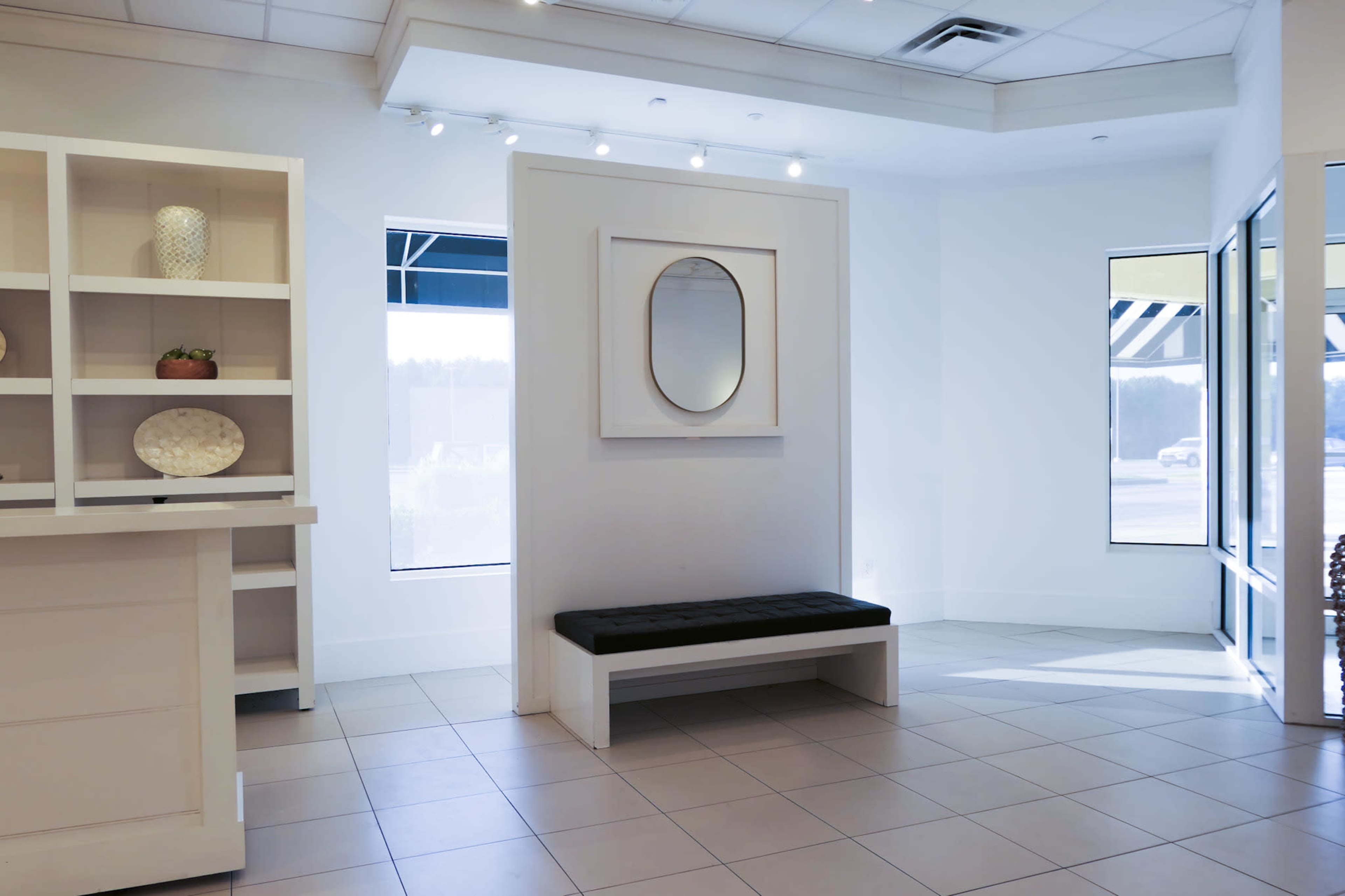The image shows a modern interior of a lobby or reception area featuring a bench, a mirror on the wall, and large windows allowing natural light.