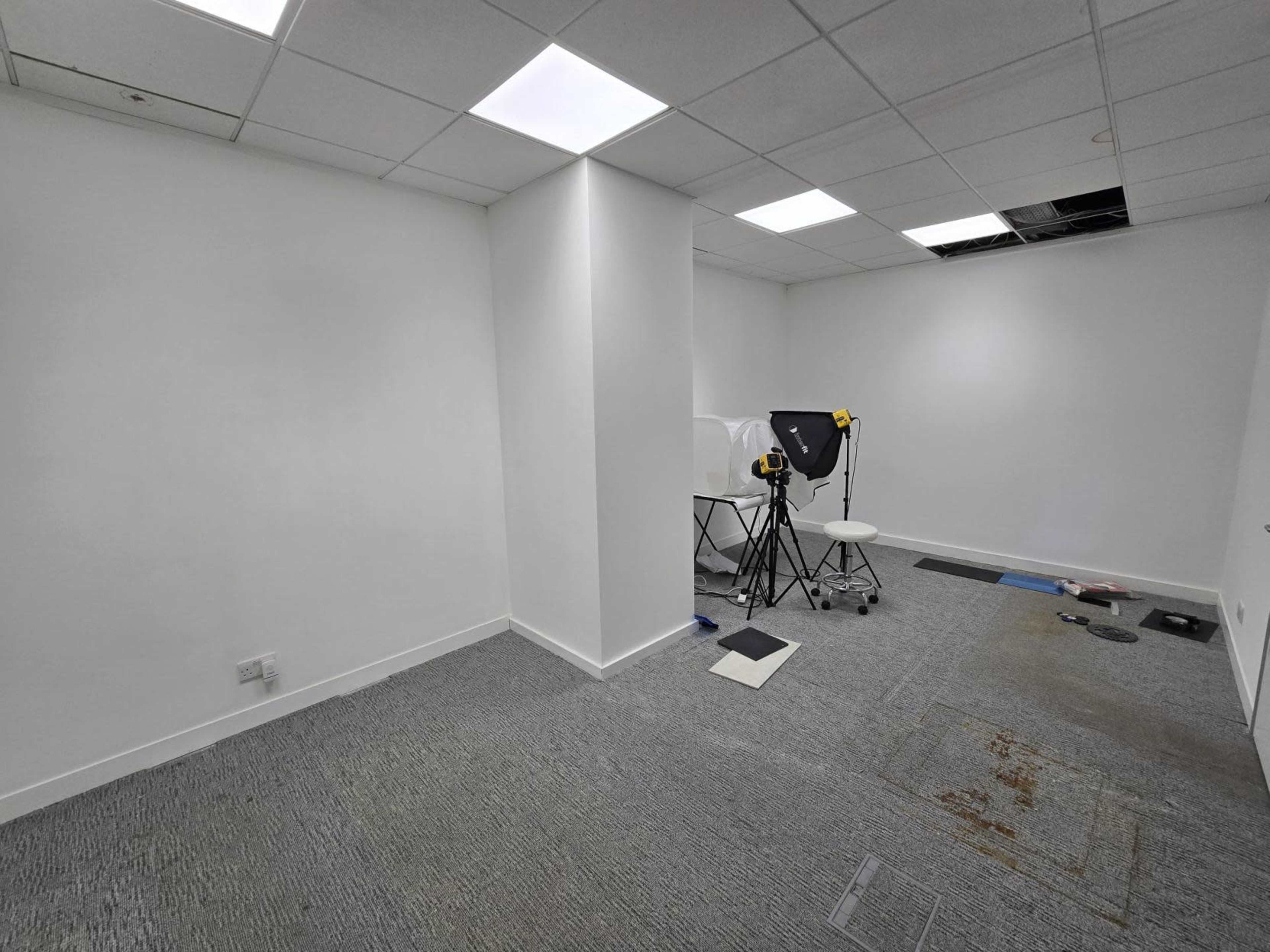 The image shows an empty room with white walls and a gray carpet, featuring a white table, two lighting stands, and a partially visible wall section with a ceiling disruption.