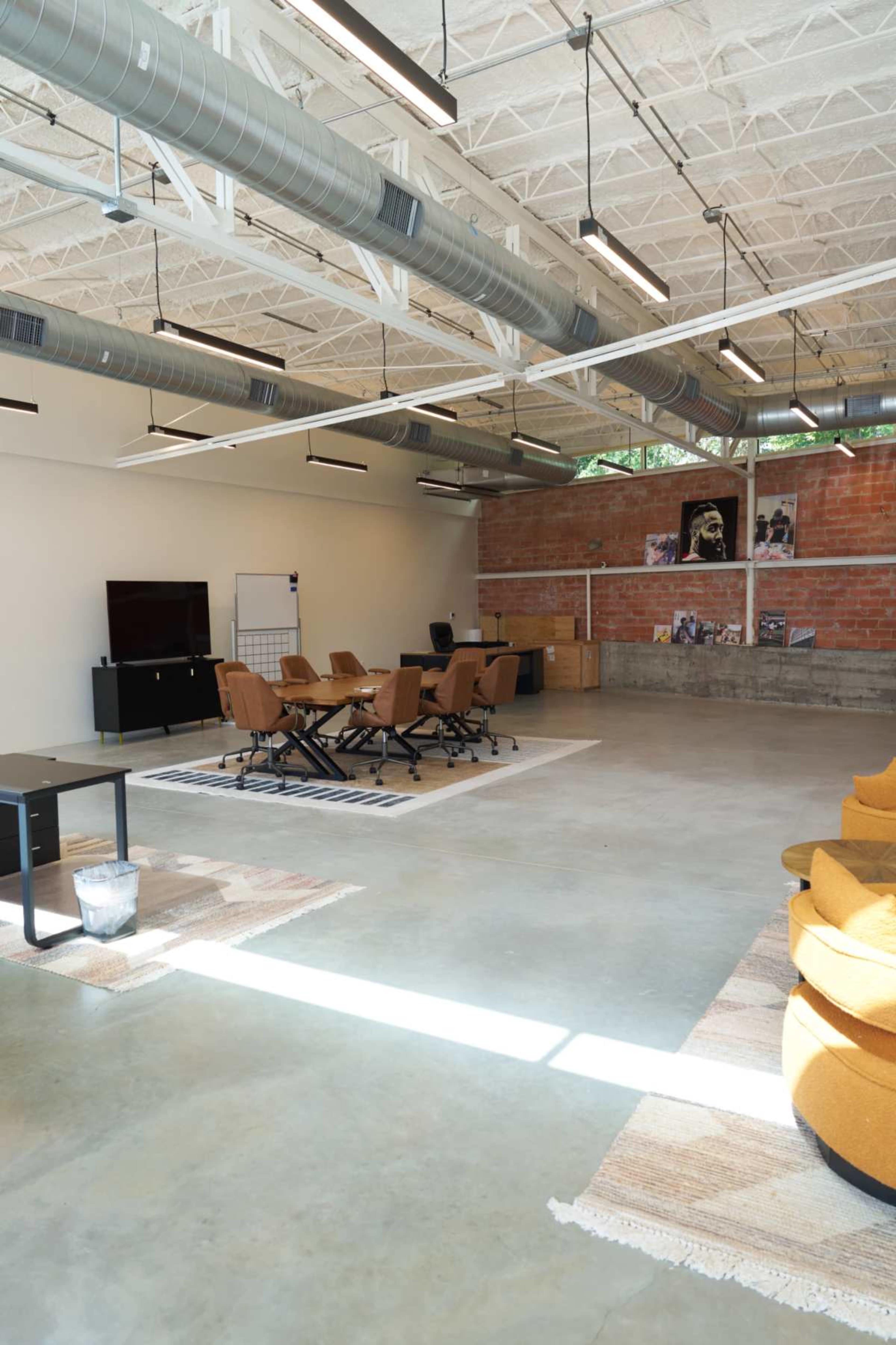 The image shows a spacious office area with a seating arrangement of brown chairs around a table, a large TV screen, and exposed ductwork on the ceiling.