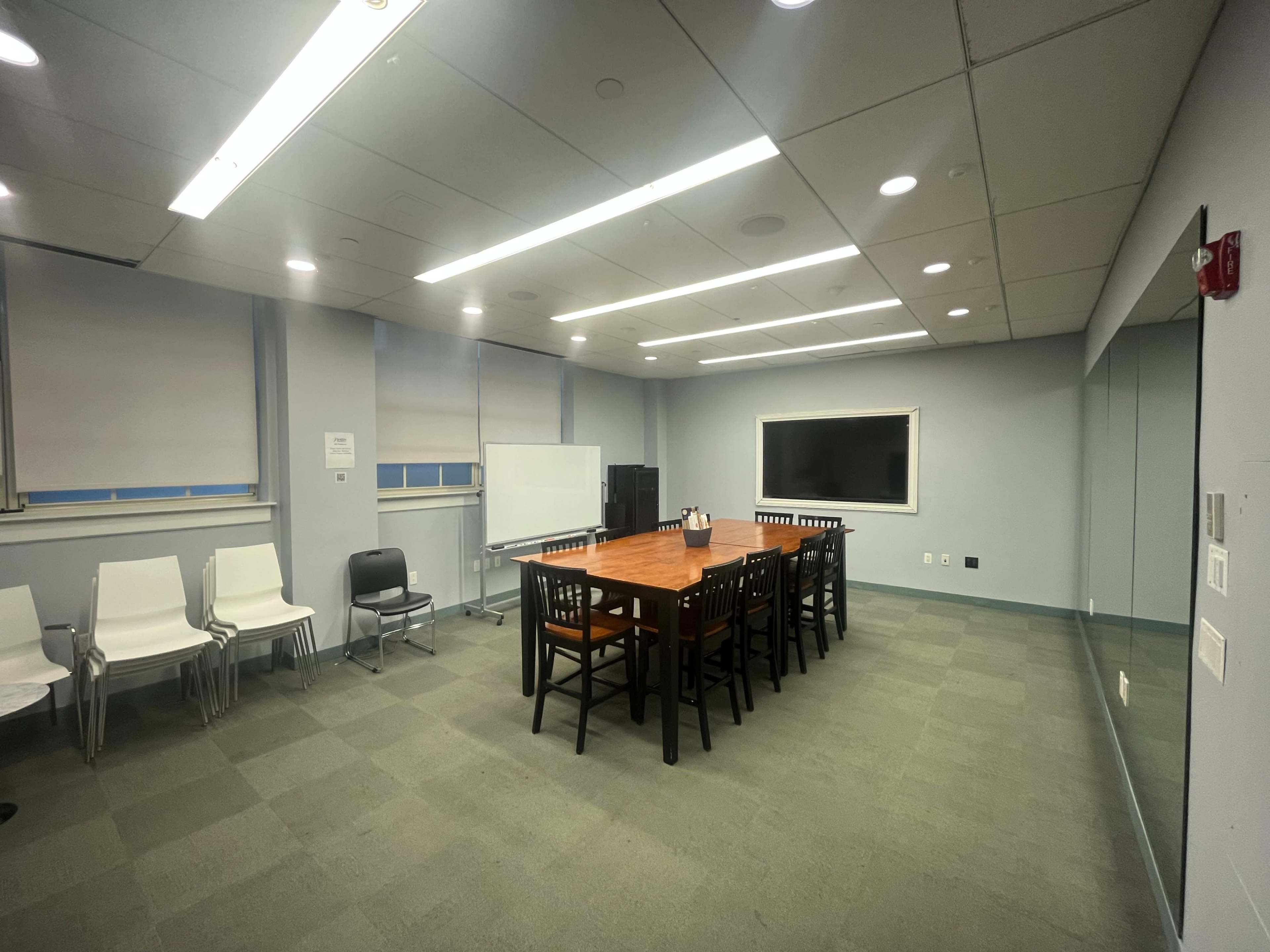 The image shows a conference room with a long wooden table, surrounded by chairs, and a large screen on one wall.