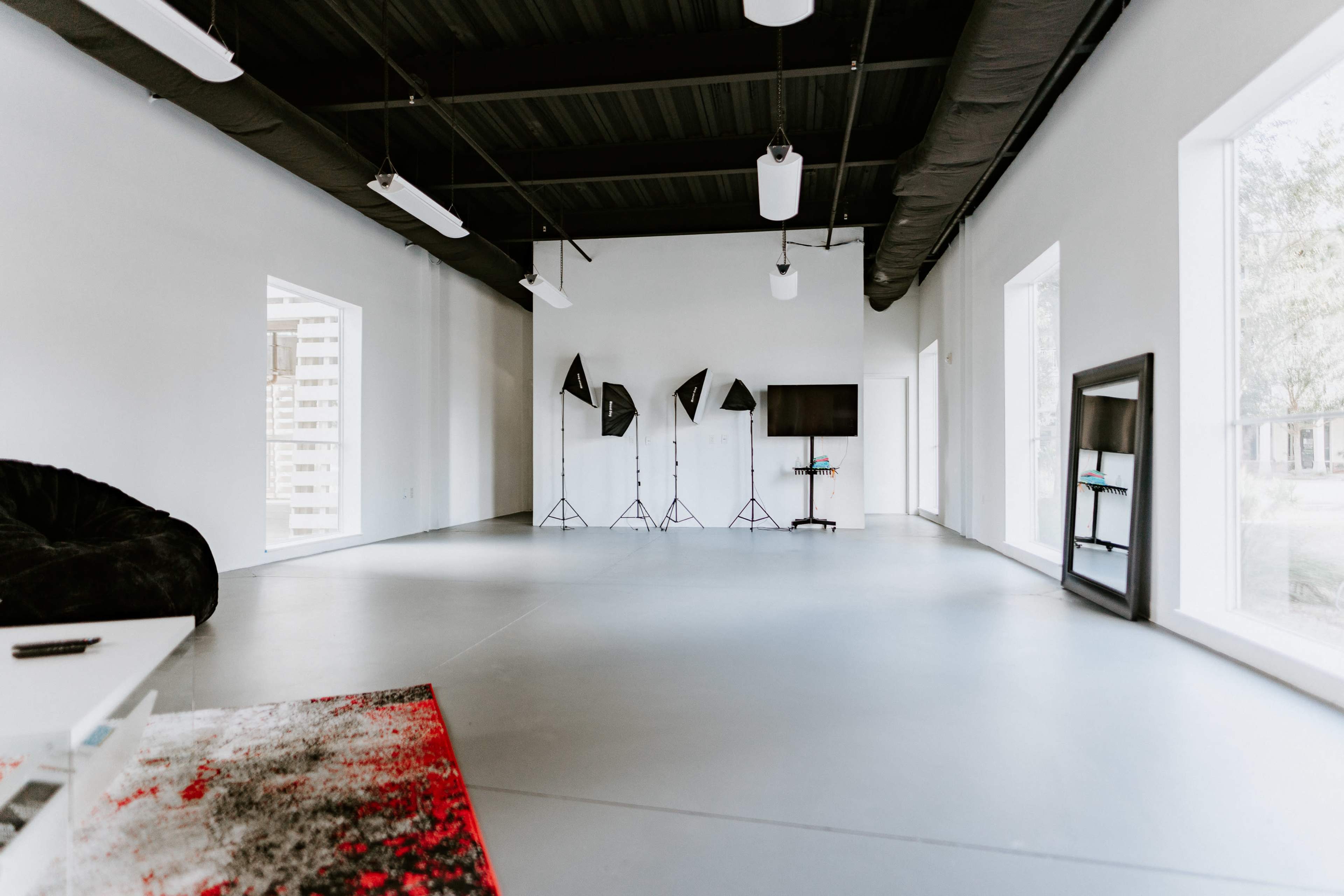 A spacious, minimally furnished studio with large windows, three lighting stands, and a television mounted on a wall.