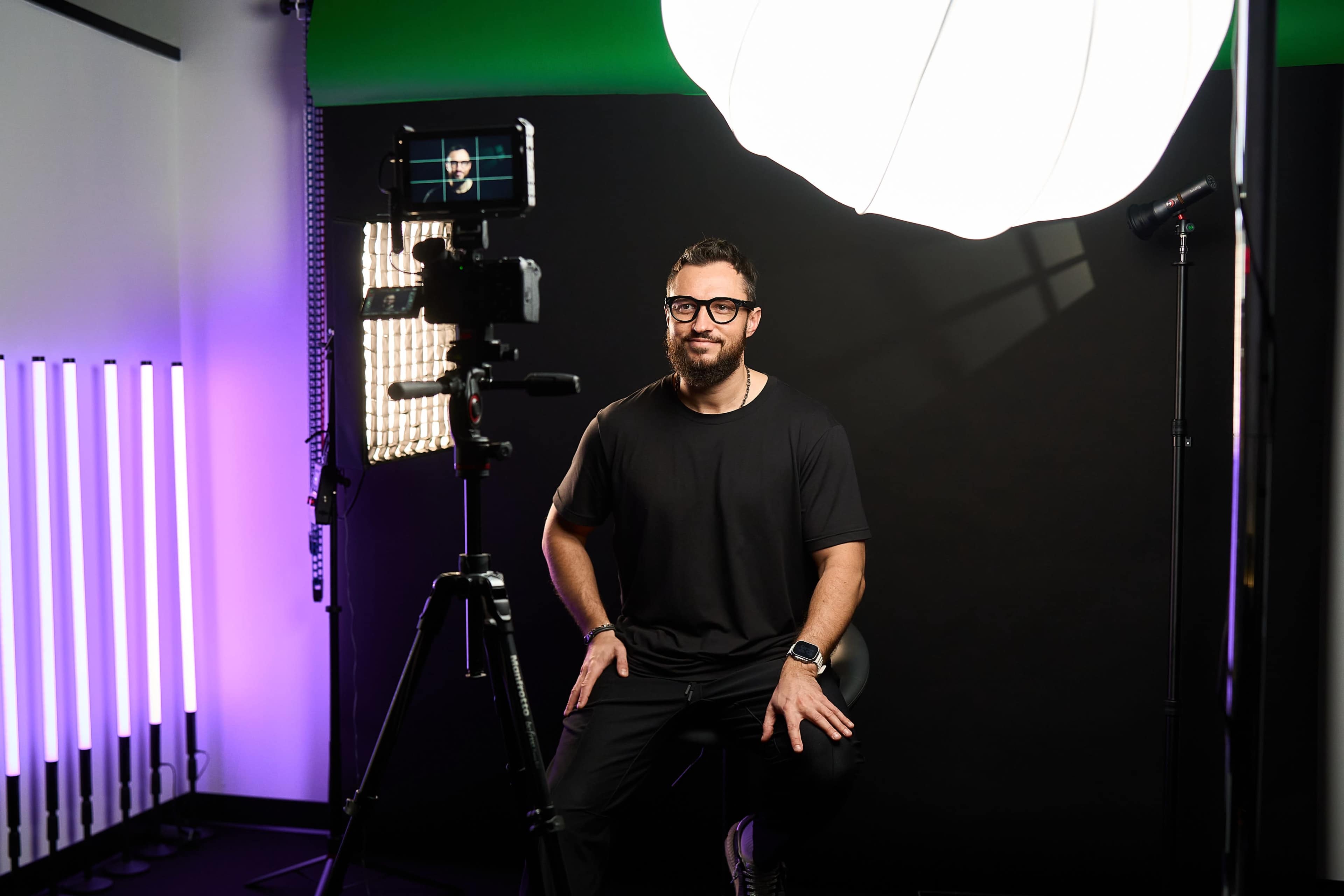A man in a black shirt sits on a stool in a studio setting, facing a camera with multiple lights and a green screen backdrop.